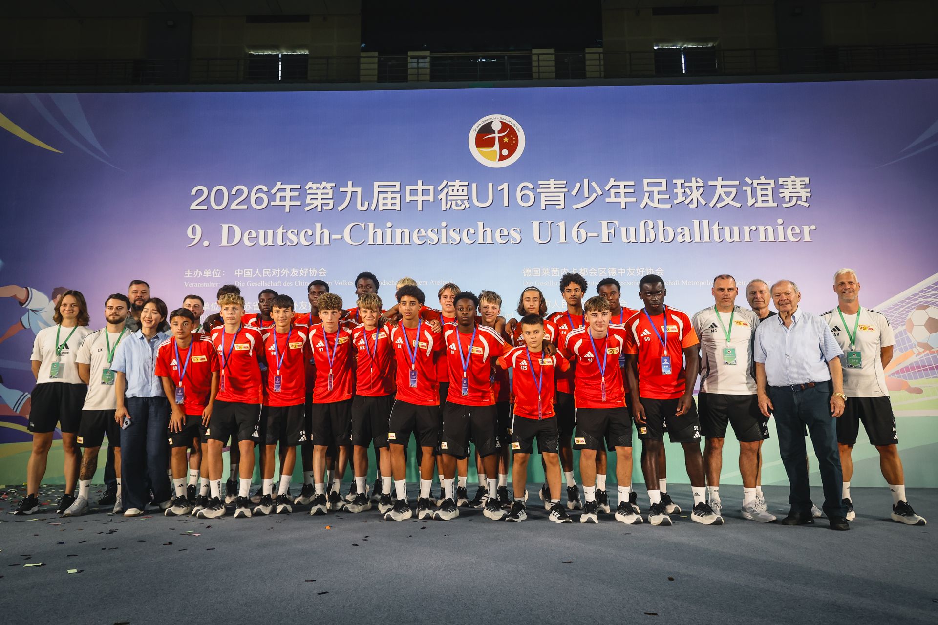 Gruppenfoto einer Fußballmannschaft mit Spielern in roten Trikots, umgeben von Trainern und Betreuern bei einem U16-Turnier.