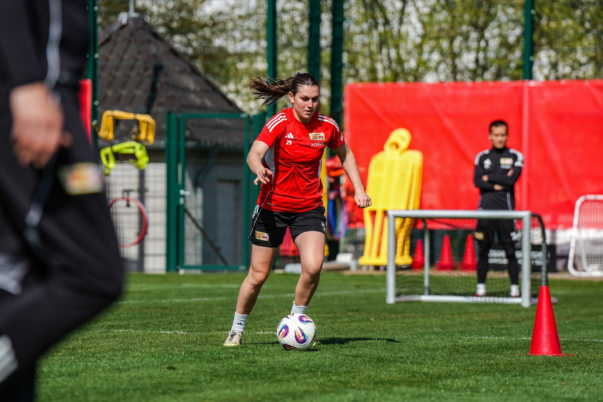 Eine Fußballspielerin in roter Trainingskleidung dribbelt einen Ball auf einem Sportplatz, während Trainer beobachten.