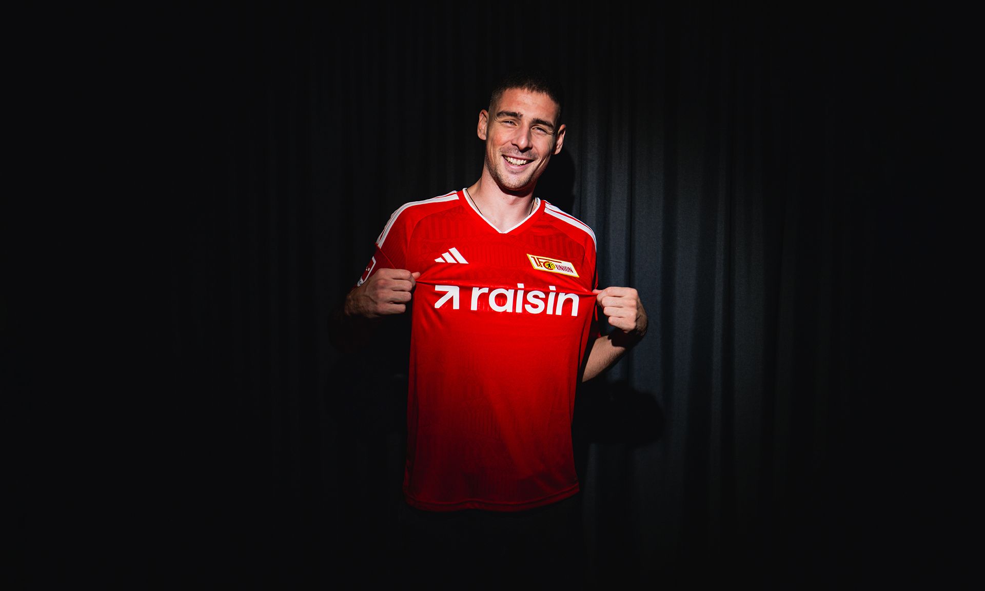 Ein Fußballspieler in einem roten Trikot mit dem Schriftzug "raisin" lächelt und hält sein Trikot hoch.