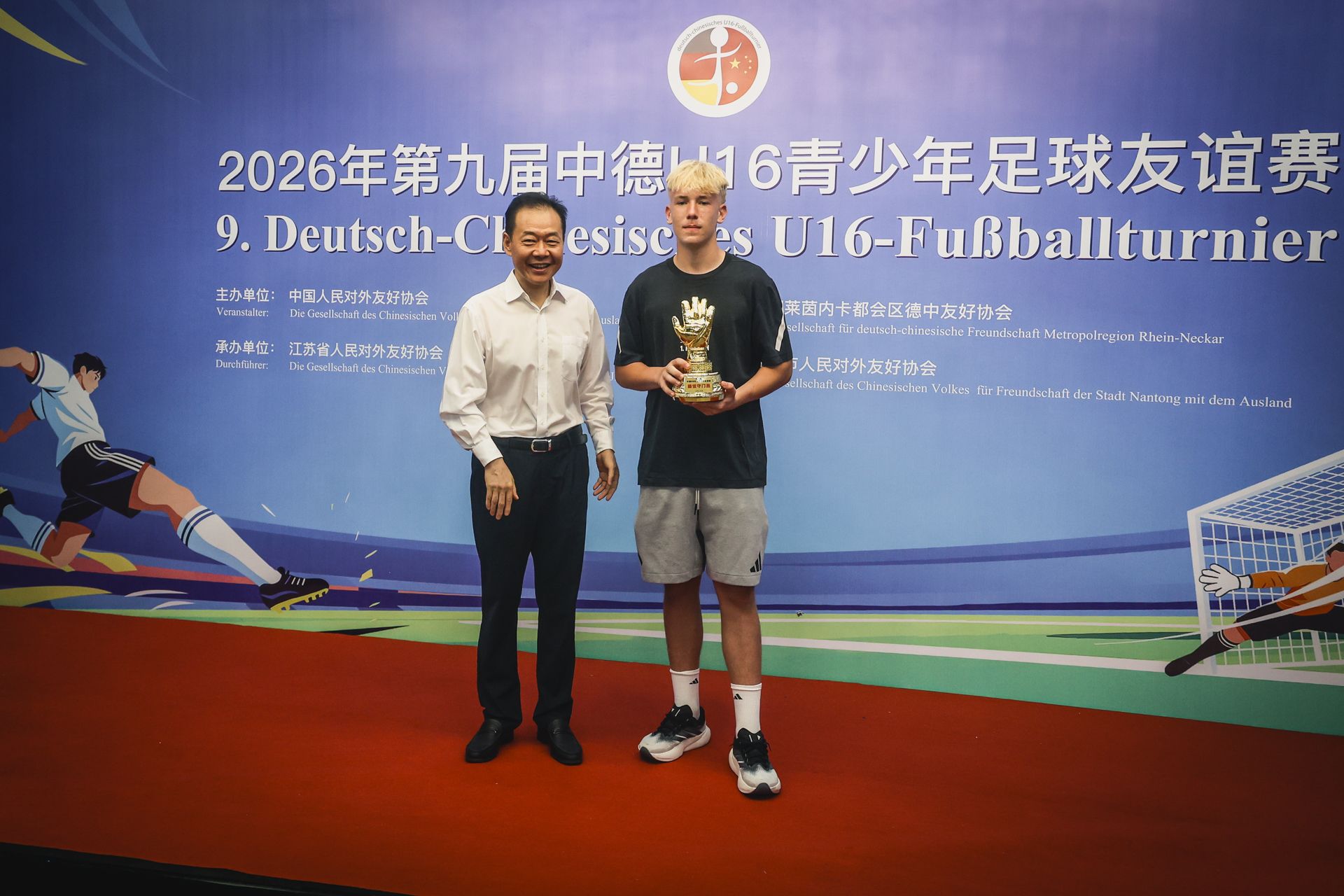 Ein junger Fußballspieler hält einen Pokal, während er neben einem älteren Mann vor einer Wand mit Turnierinformationen steht.