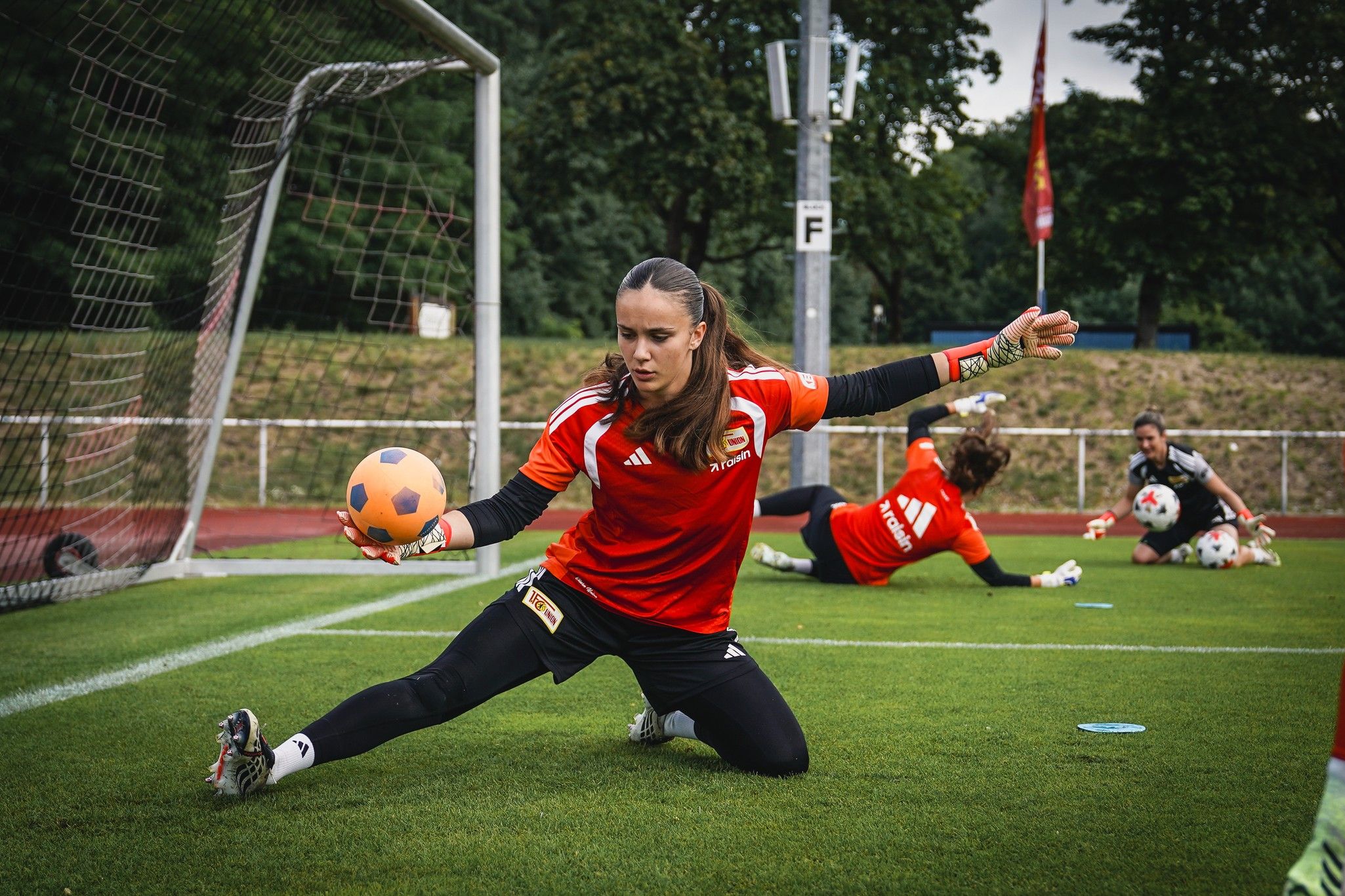 Eine Torhüterin in rotem Trikot fängt einen Ball im Training auf dem Fußballfeld, während eine weitere Spielerin im Hintergrund trainiert.