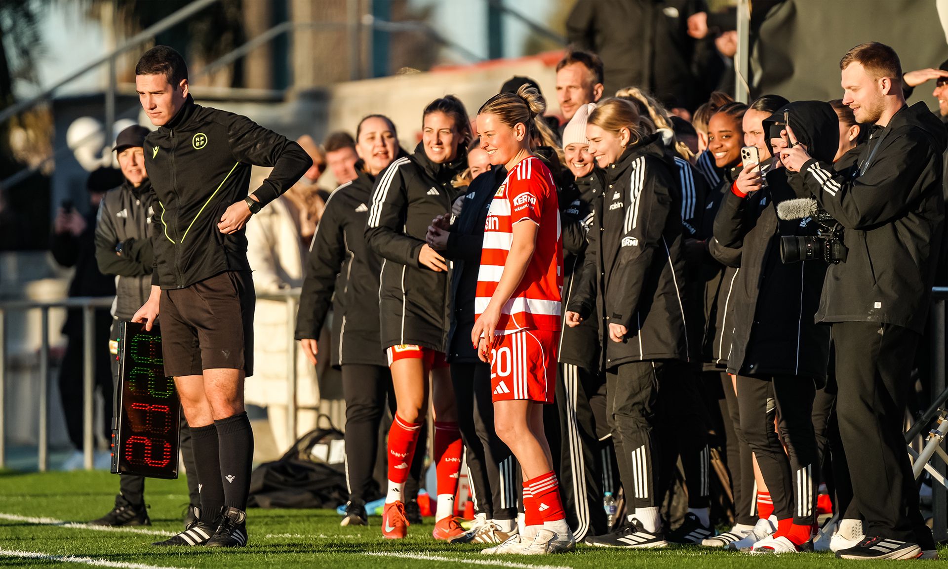 Eine Spielerin steht am Spielfeldrand, bereit zum Einwechseln, umgeben von lächelnden Teamkollegen und einem Schiedsrichter.