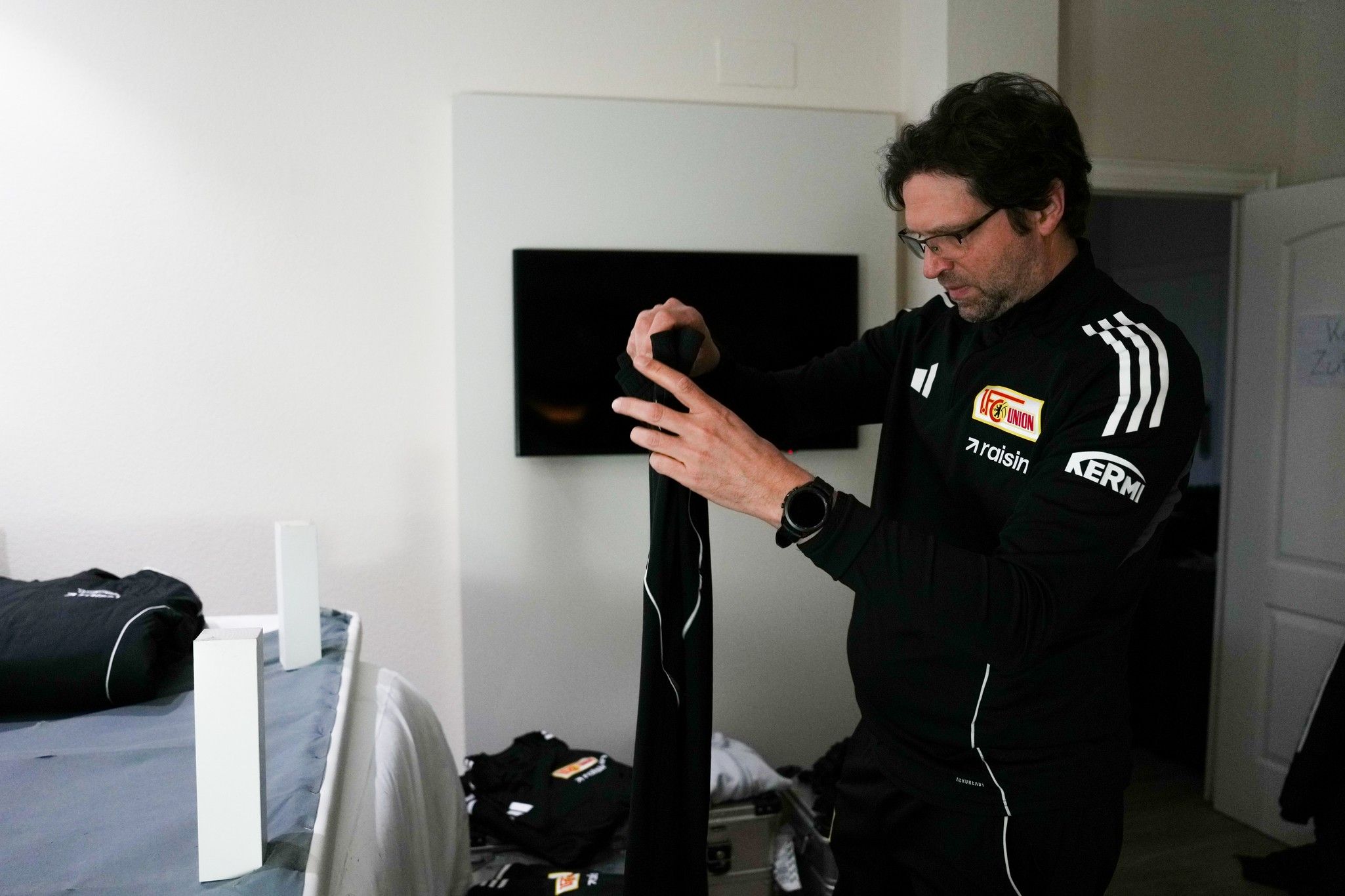 Ein Mann in Sportbekleidung faltet einen schwarzen Kleidungsstück in einem Raum mit Fernseher und Koffer.