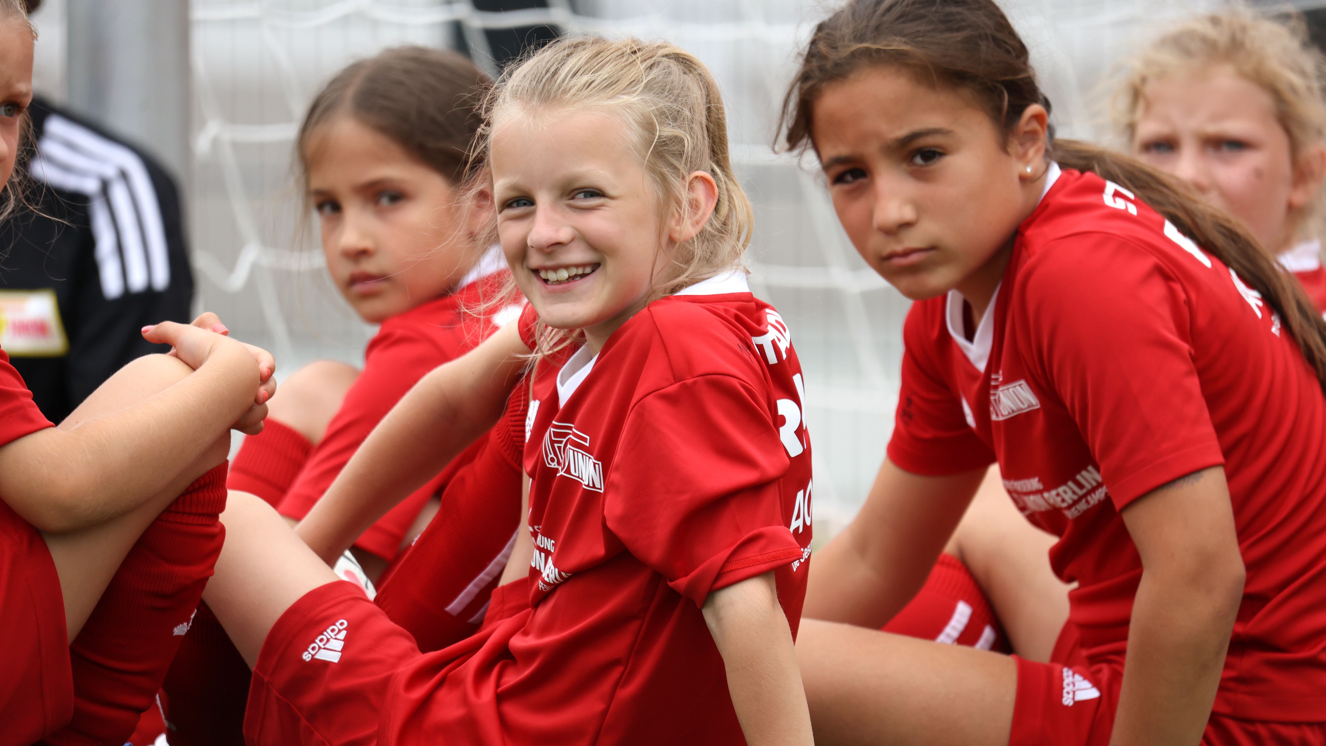 Drei Mädchen in roten Fußballtrikots sitzen zusammen und blicken in die Kamera, während sie auf dem Spielfeld entspannen.