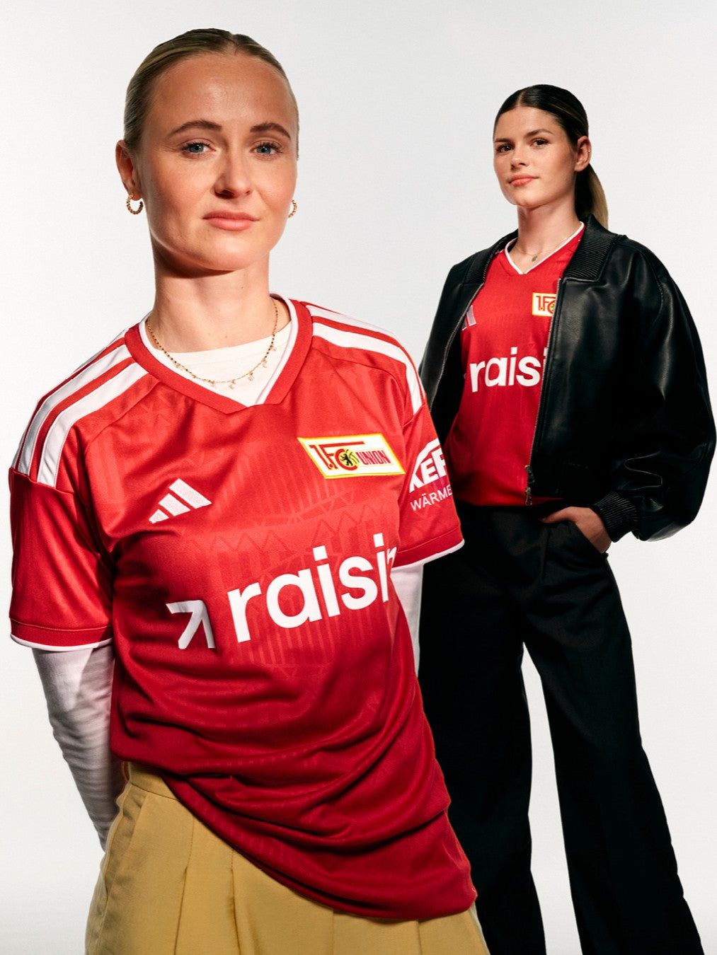 Zwei Frauen in roten Fußballtrikots stehen vor einem weißen Hintergrund, eine mit lässiger Kleidung und die andere mit sportlichem Outfit.