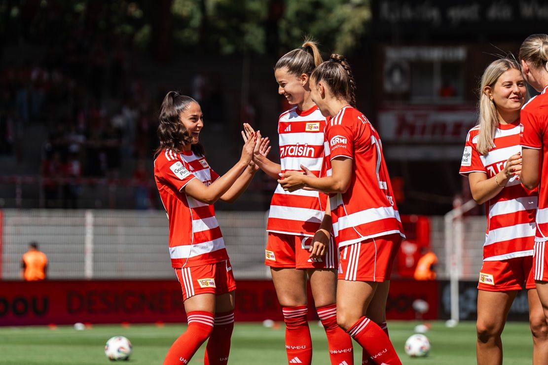 Vier Fußballspielerinnen in roten Trikots stehen auf dem Spielfeld und feiern miteinander. Im Hintergrund Zuschauer und Stadionatmosphäre.