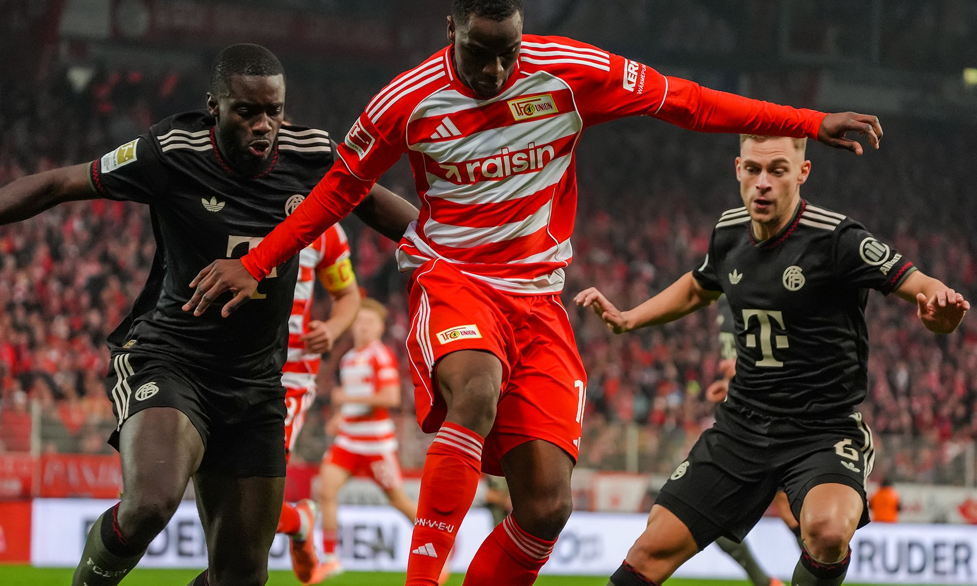 Fußballspiel mit einem Spieler in rotem Trikot, der von zwei Spielern in schwarzen Trikots bedrängt wird. Stadionatmosphäre.