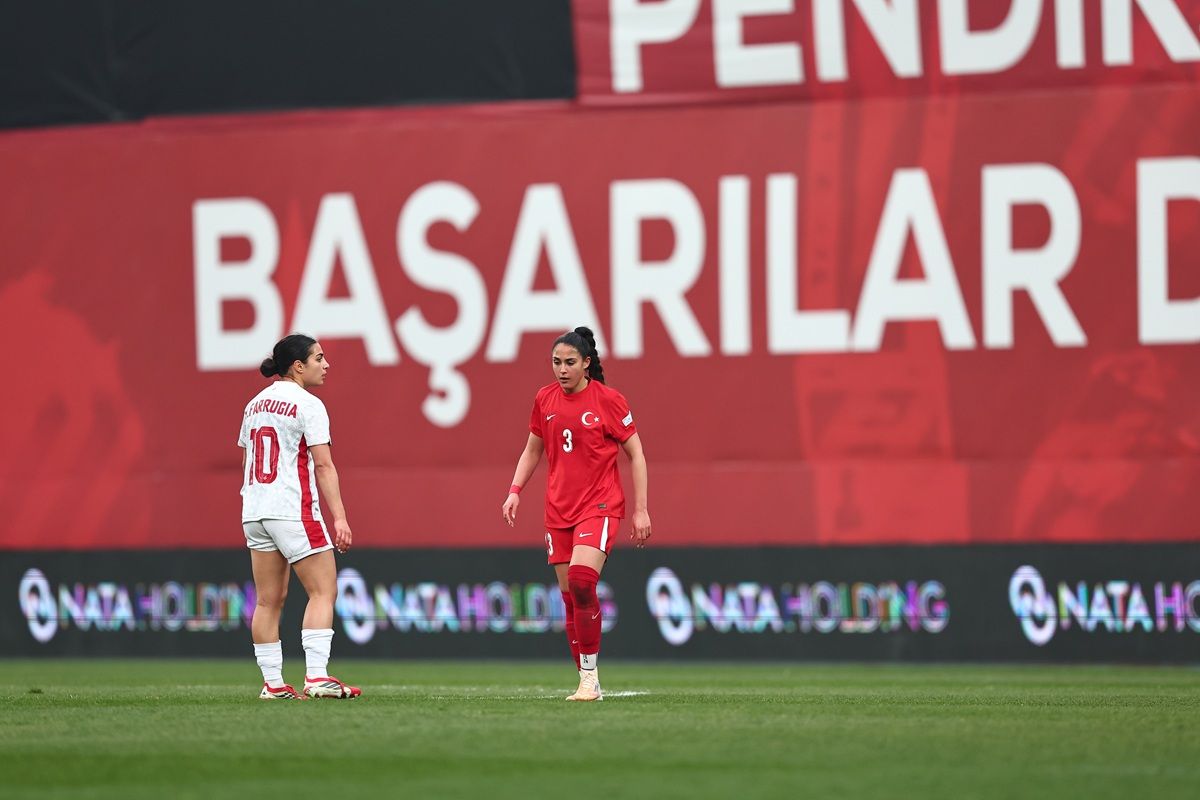 Zwei Fußballspielerinnen auf dem Spielfeld, eine in roter und die andere in weißer Uniform, vor einem großen Werbebanner.
