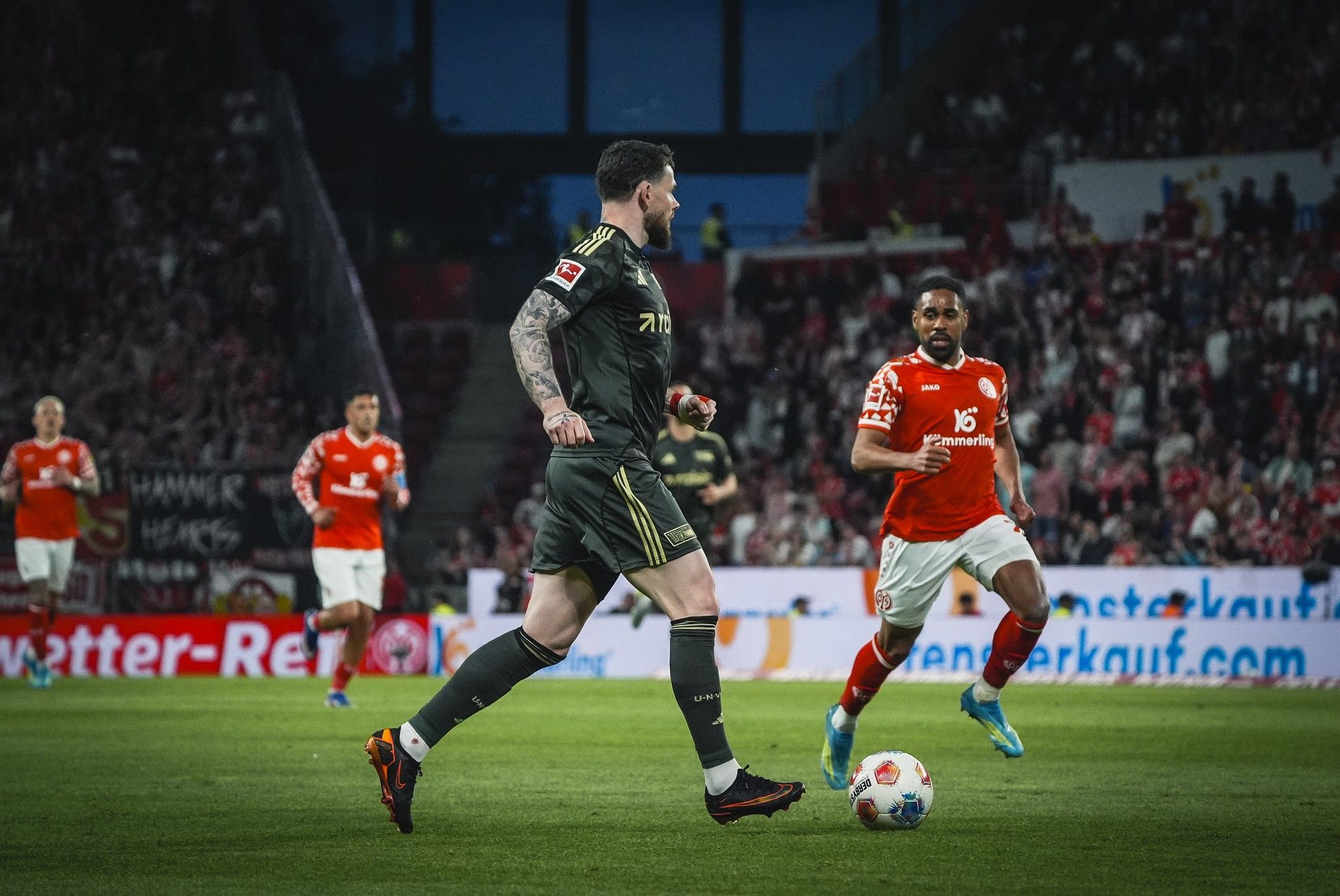 Fußballspiel mit Spielern in schwarzen und roten Trikots auf dem Platz, Zuschauermenge im Hintergrund.