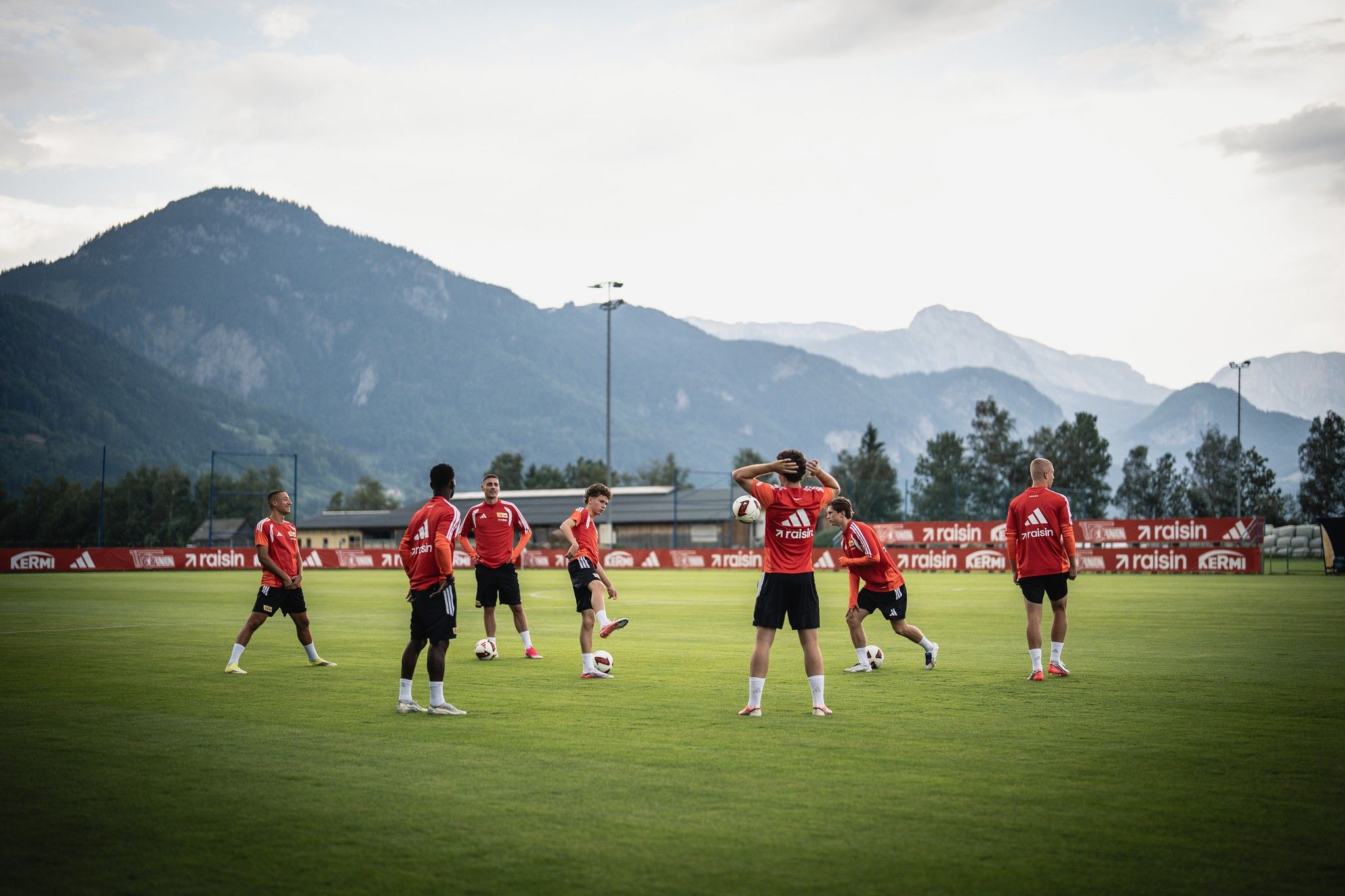 Fußballmannschaft in roten Trikots trainiert auf einem Rasenplatz mit Bergkulisse im Hintergrund.