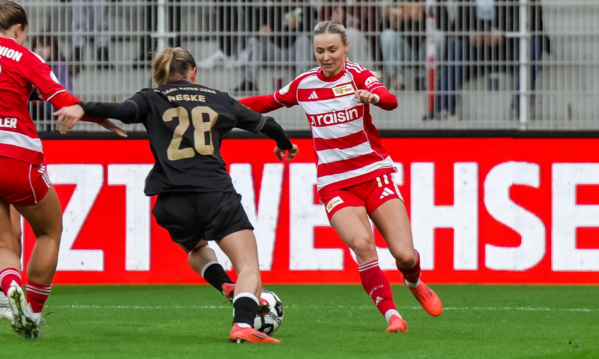 Eine Fußballspielerin im roten Trikot dribbelt am Gegner vorbei, während eine Torhüterin sich darauf vorbereitet, den Ball abzufangen.