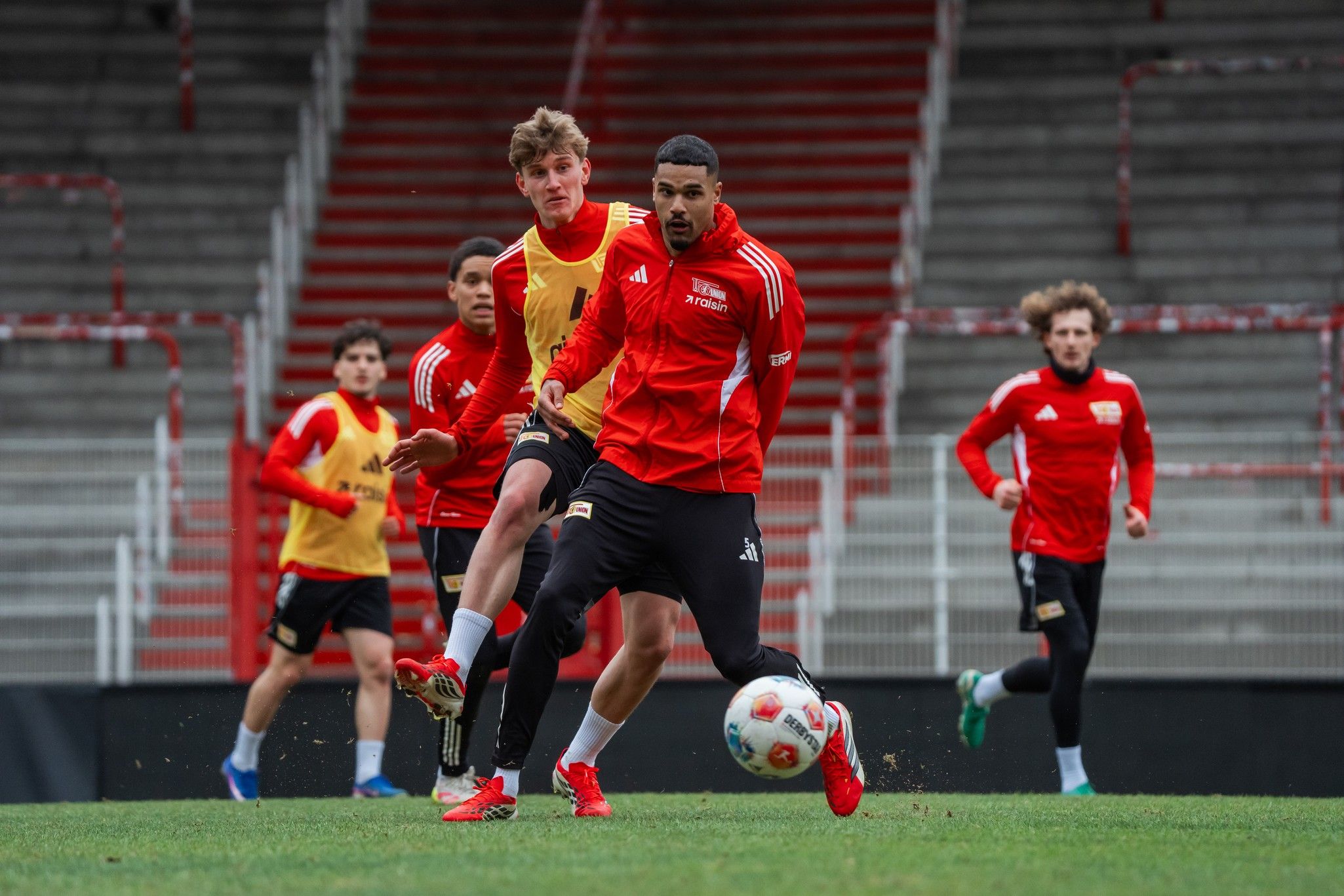 Zwei Fußballspieler in roten Trikots konkurrieren um den Ball auf einem Trainingsplatz, während weitere Spieler im Hintergrund stehen.