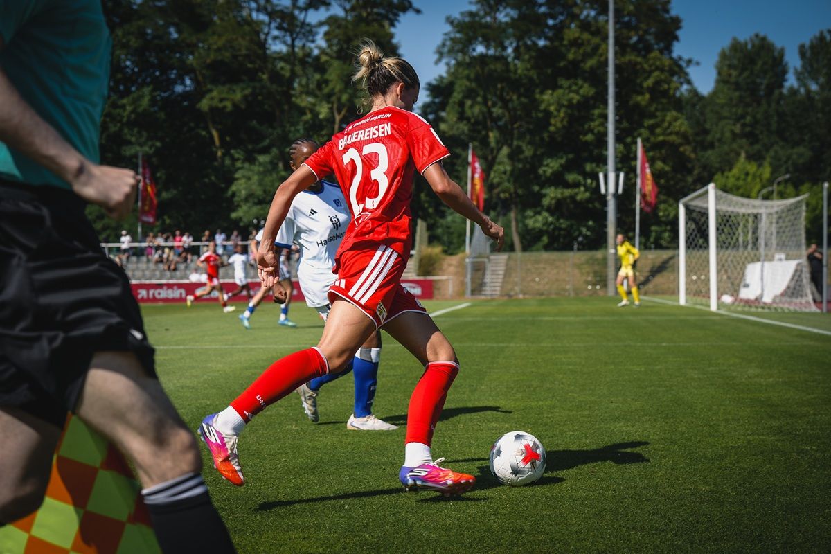Eine Spielerin in rotem Trikot mit der Nummer 23 dribbelt den Ball auf einem Fußballfeld, während ein Gegner in weißem Trikot verfolgt.