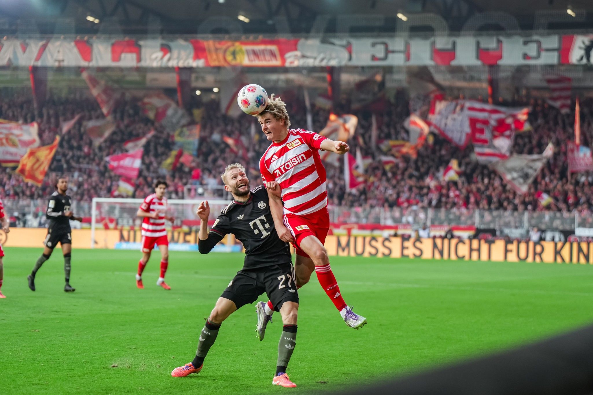 Ein Fußballspieler springt hoch, um den Ball zu köpfen, während ein Gegner ihn herausfordert. Im Hintergrund Fans mit Flaggen.