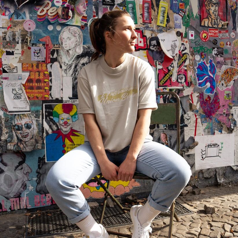 Eine Person sitzt auf einem Stuhl vor einer bunten Wand aus Plakaten und Graffiti, trägt ein helles T-Shirt und Jeans.