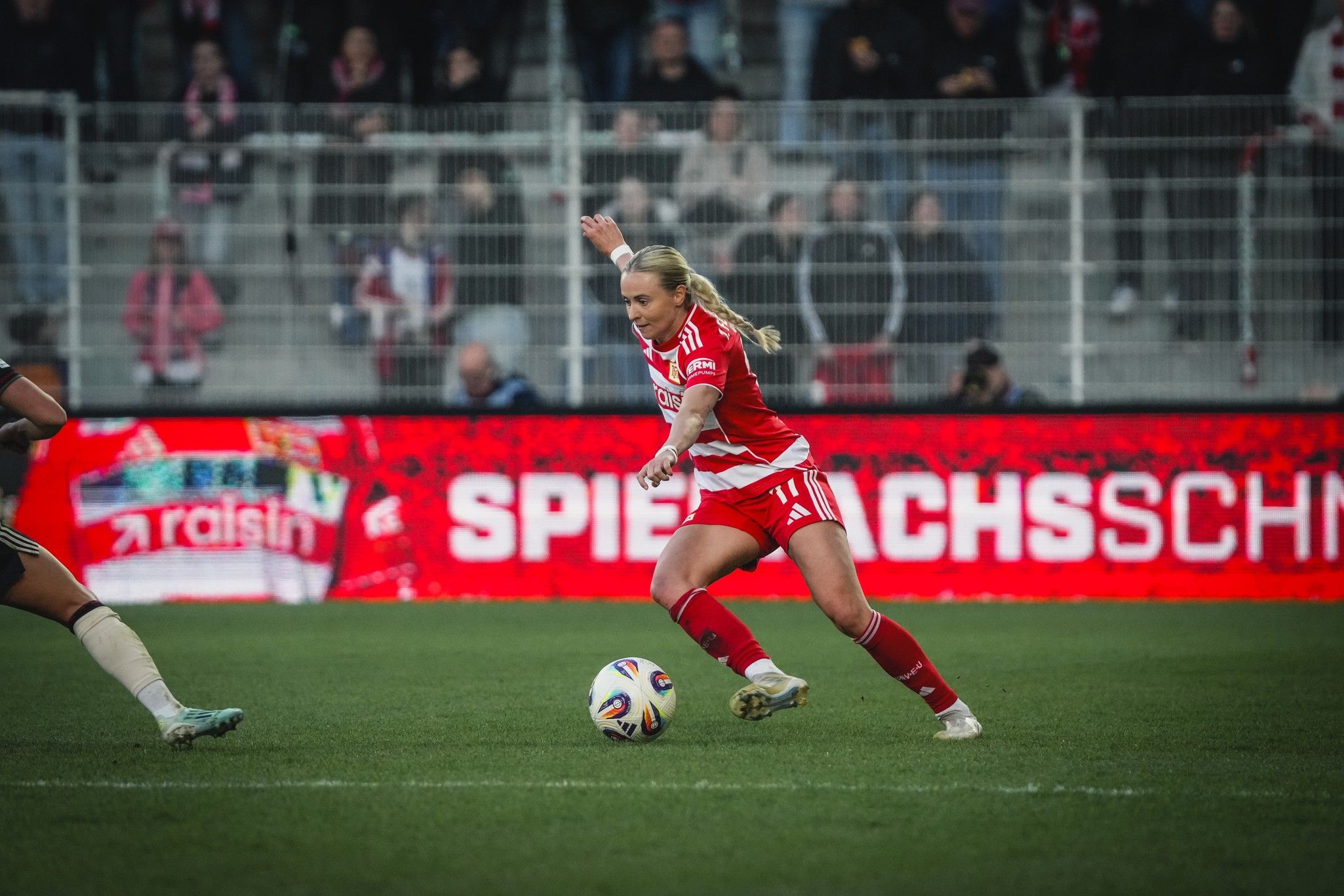Eine Fußballspielerin dribbelt den Ball auf einem Spielfeld, im Hintergrund sind Zuschauer und Werbung zu sehen.