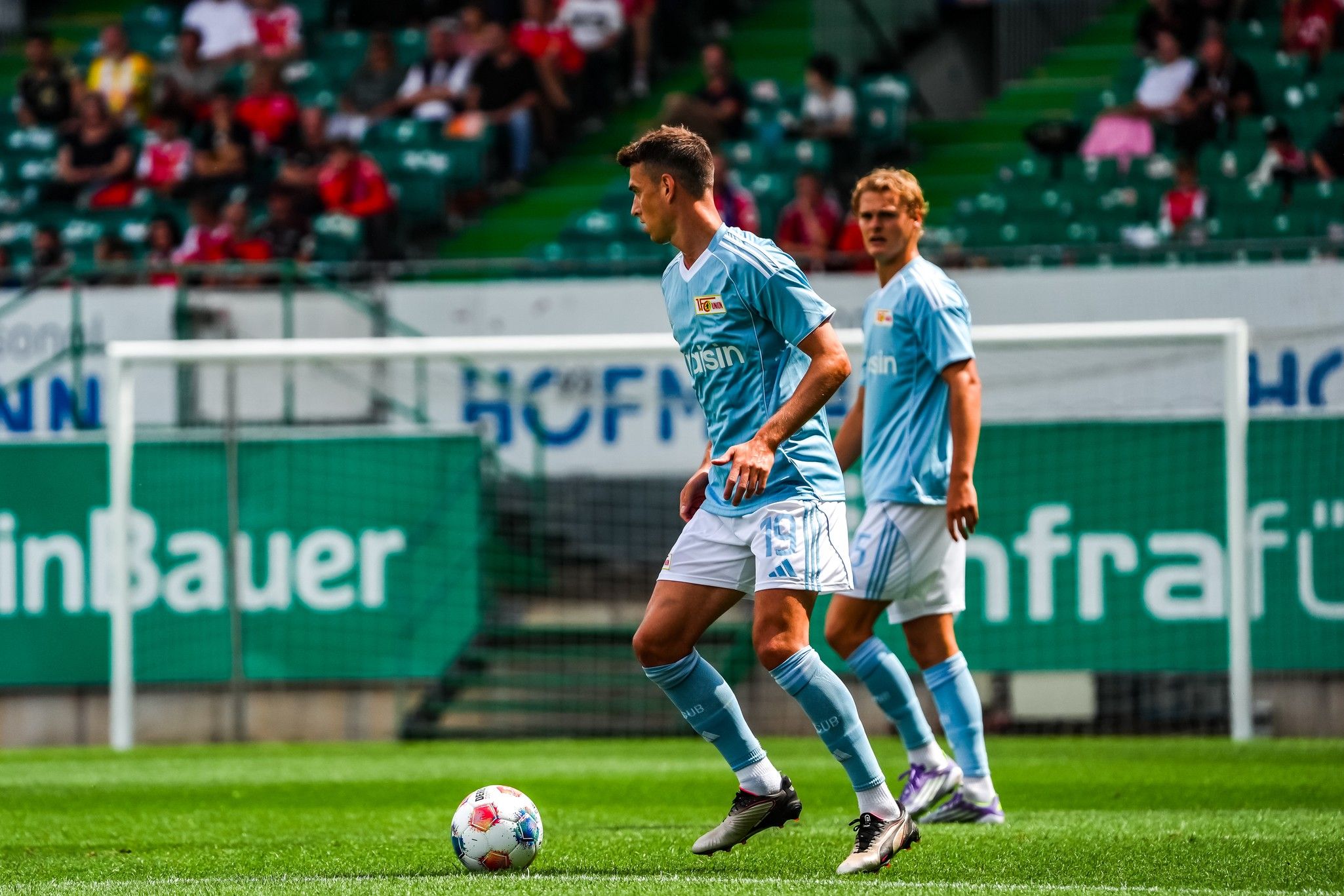 Zwei Fußballspieler in blauen Trikots auf dem Spielfeld, im Hintergrund eine Zuschauertribüne.
