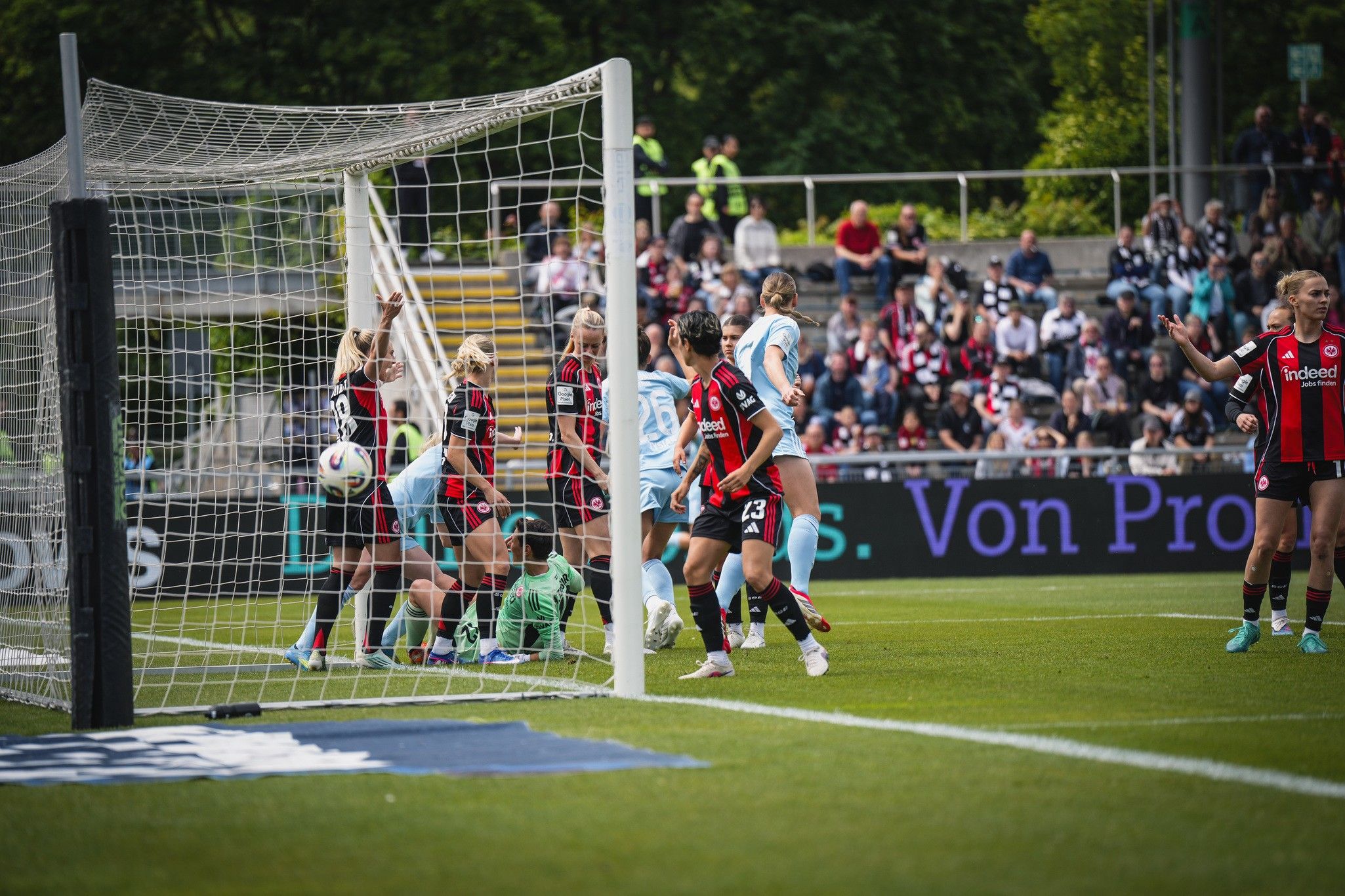 Spielszene im Frauenfußball mit mehreren Spielerinnen in einem Torraum, während ein Ball ins Netz fliegt. Zuschauer im Hintergrund.