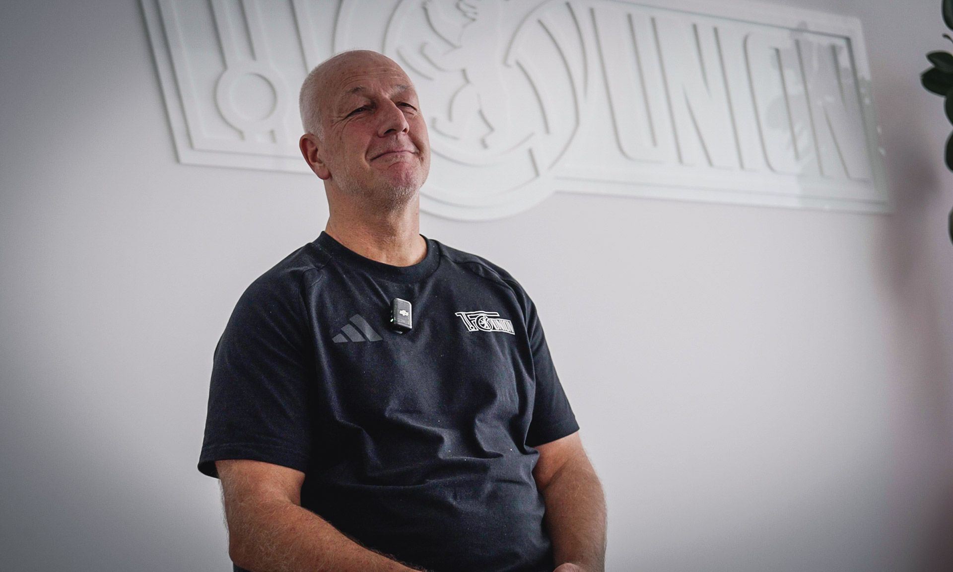 Ein lächelnder Mann sitzt vor einer Wand mit einem Relief des Logos "UNION". Er trägt ein schwarzes T-Shirt und hat ein Mikrofon am Oberteil.
