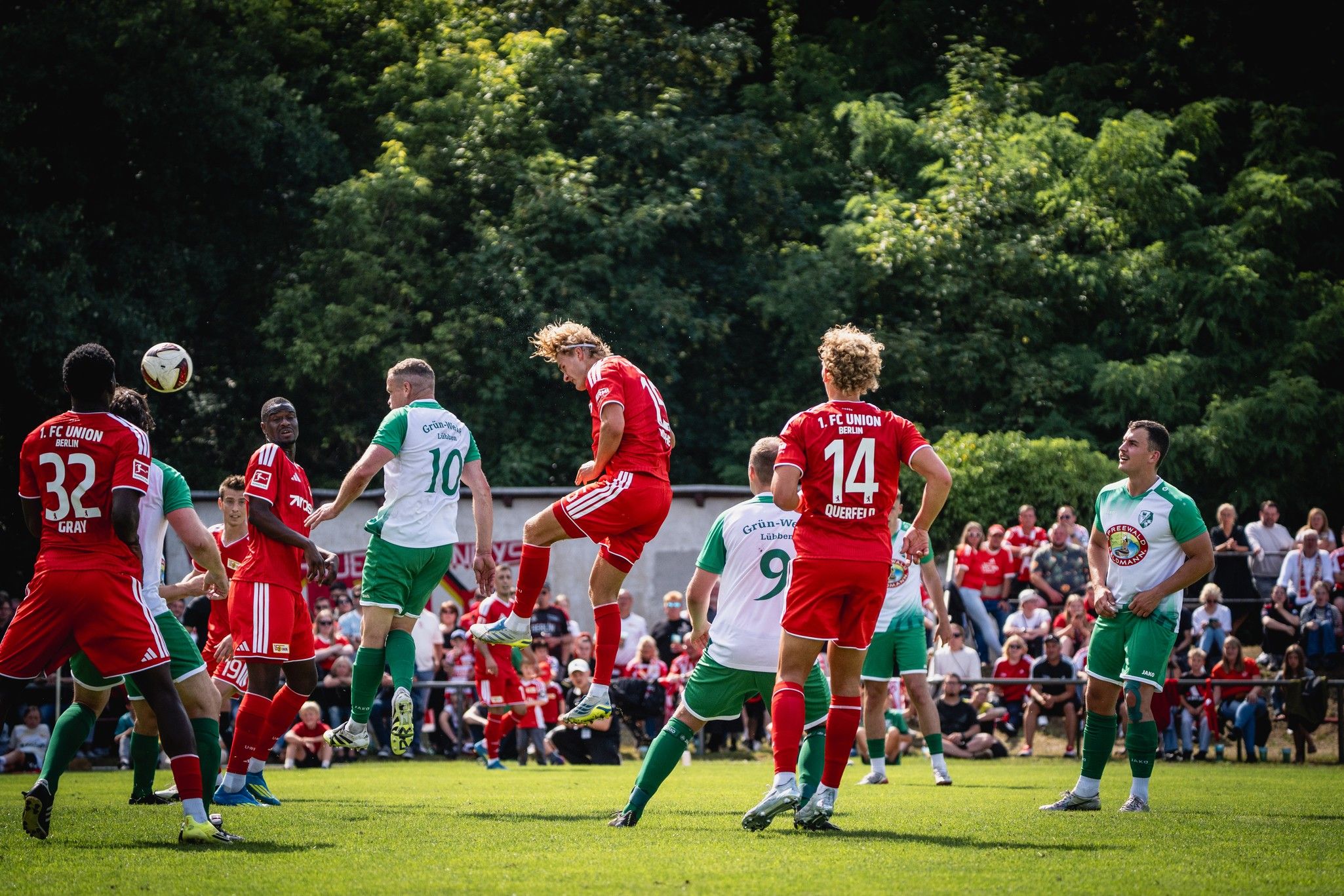 Spielszene im Fußball mit Spielern in roten und grünen Trikots, die um den Ball in einem spannenden Moment kämpfen. Zuschauer im Hintergrund.