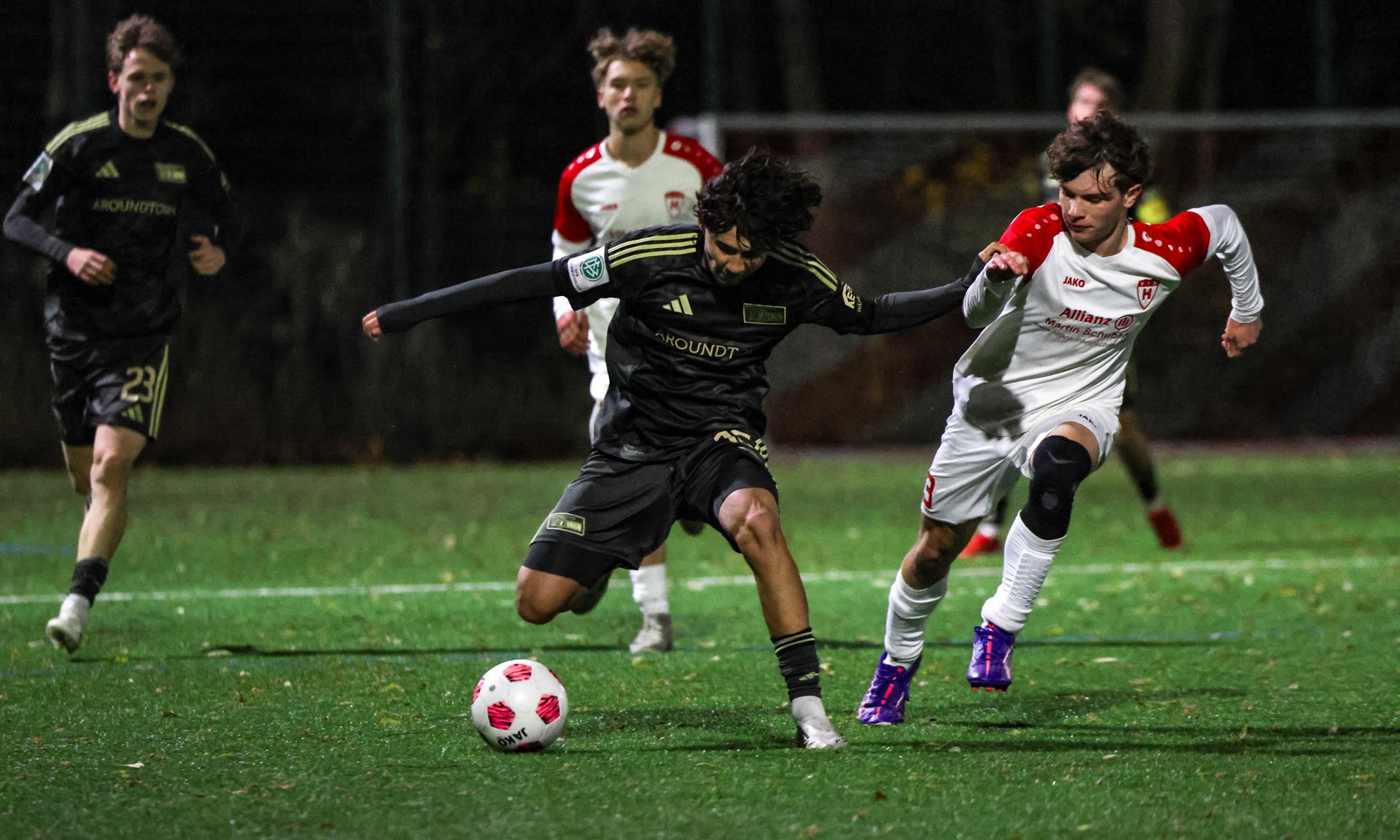 Spielszene eines Fußballspiels bei Nacht, zwei Spieler kämpfen um den Ball, im Hintergrund weitere Spieler.