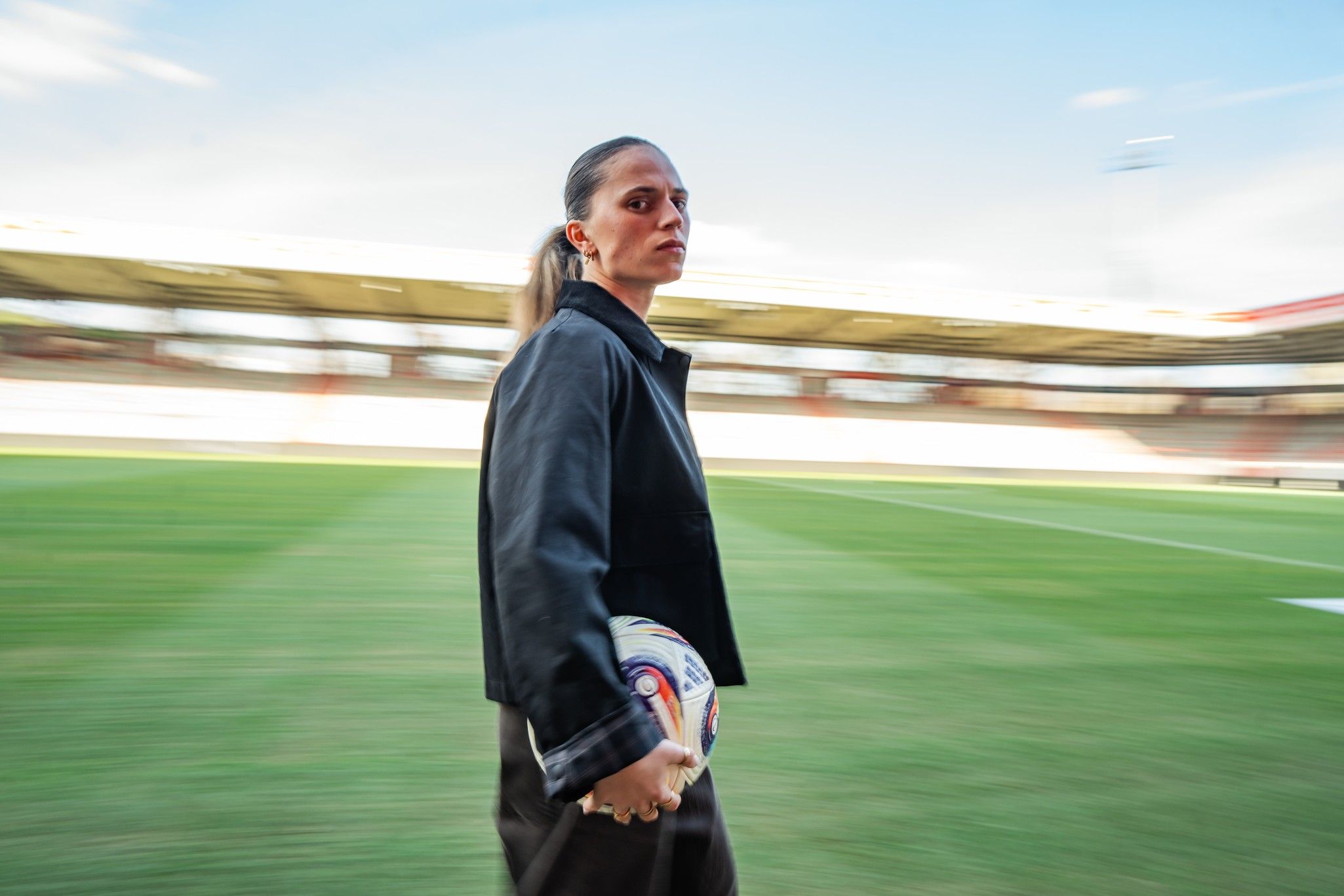 Eine Sportlerin hält einen Fußball und ist in Bewegung auf einem Fußballfeld, mit einem verschwommenen Hintergrund.