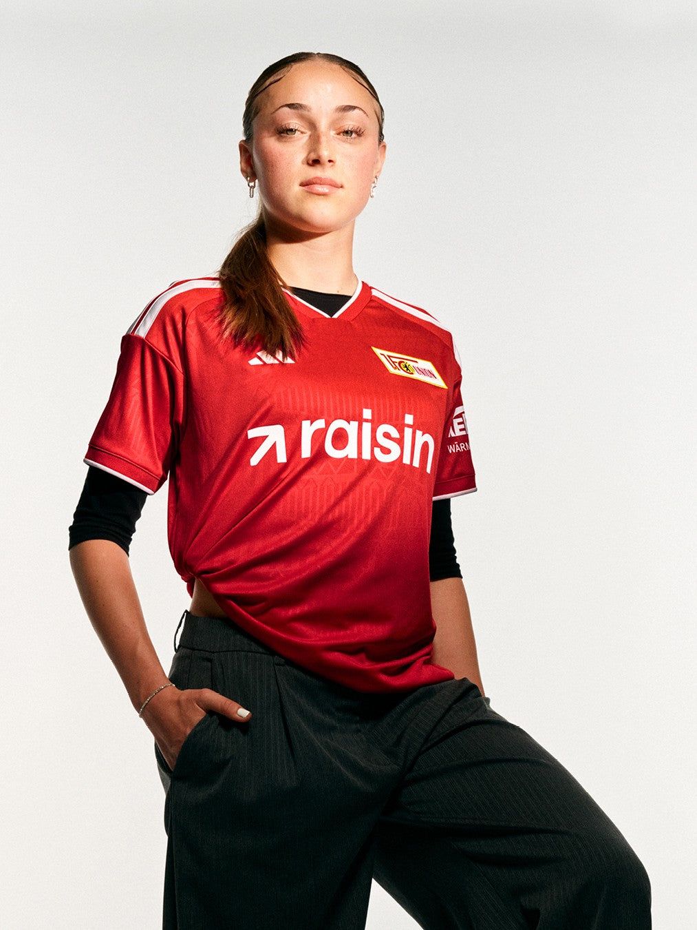 Frau in rotem Fußballtrikot mit dem Schriftzug „raisin“ und schwarzer Hose, steht selbstbewusst vor neutralem Hintergrund.