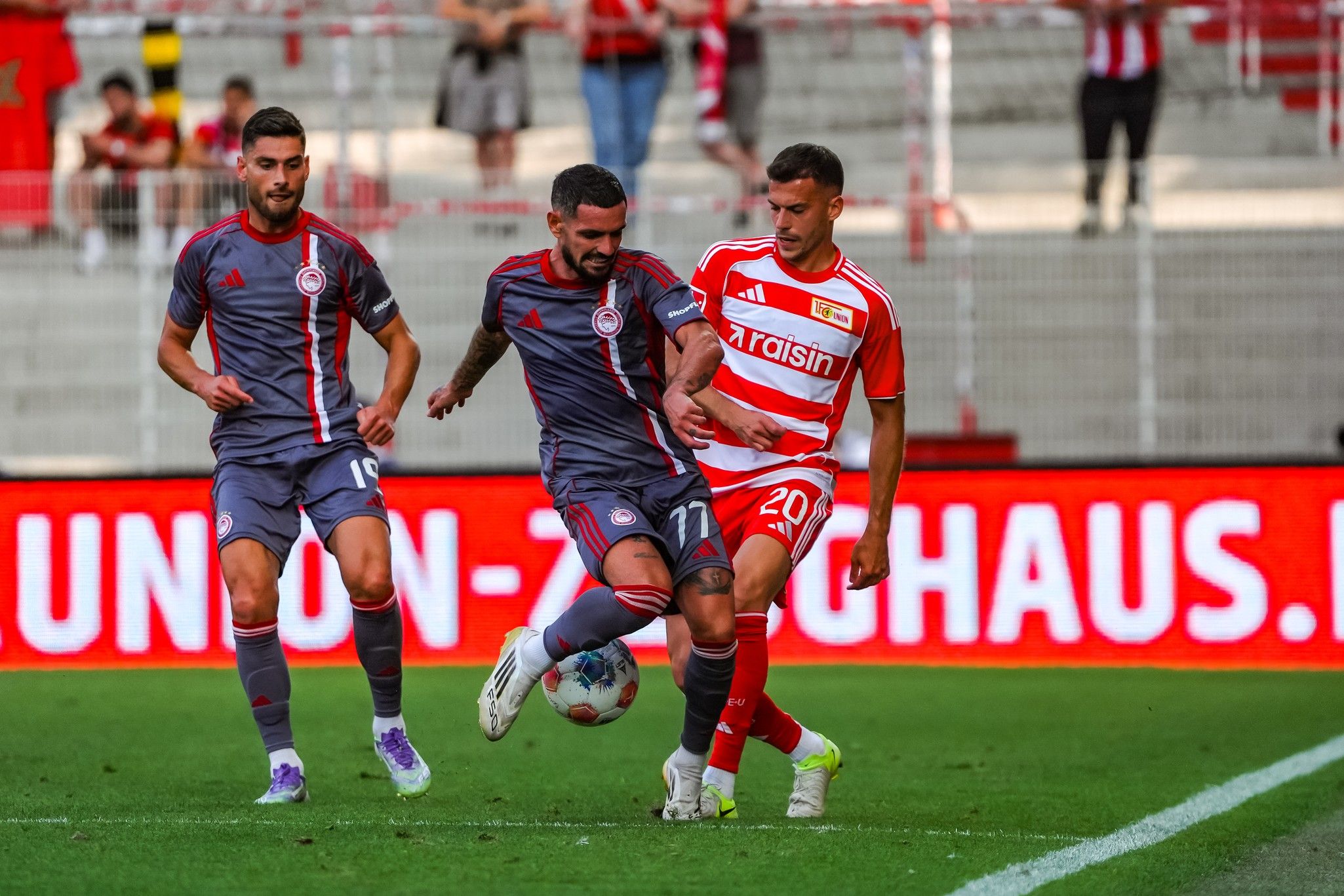Zwei Fußballspieler in grauen und roten Trikots kämpfen um den Ball auf einem Stadionfeld.