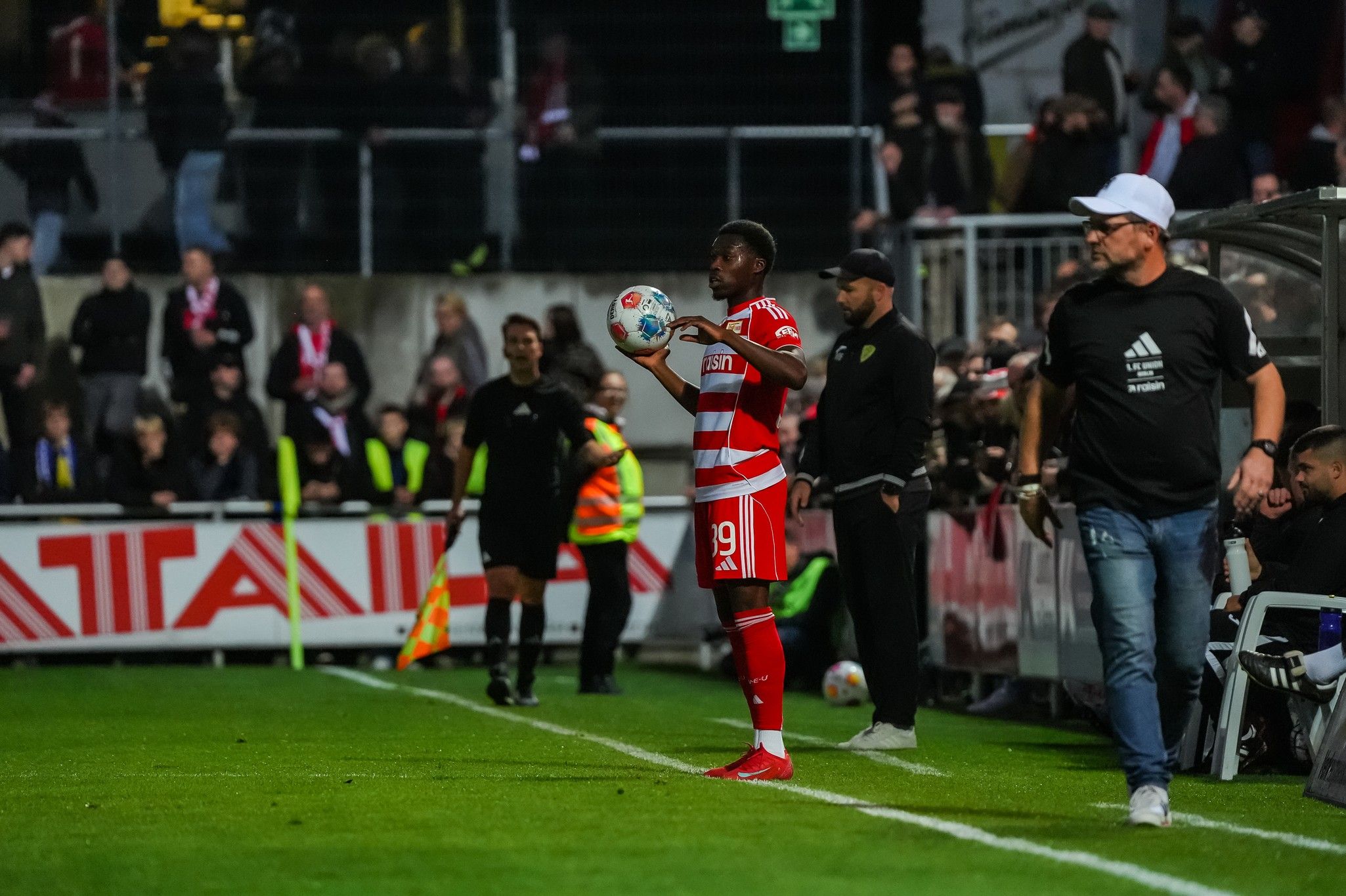 Ein Spieler in rotem Trikot bereitet sich darauf vor, einen Einwurf an der Seitenlinie auszuführen, während Zuschauer im Hintergrund stehen.