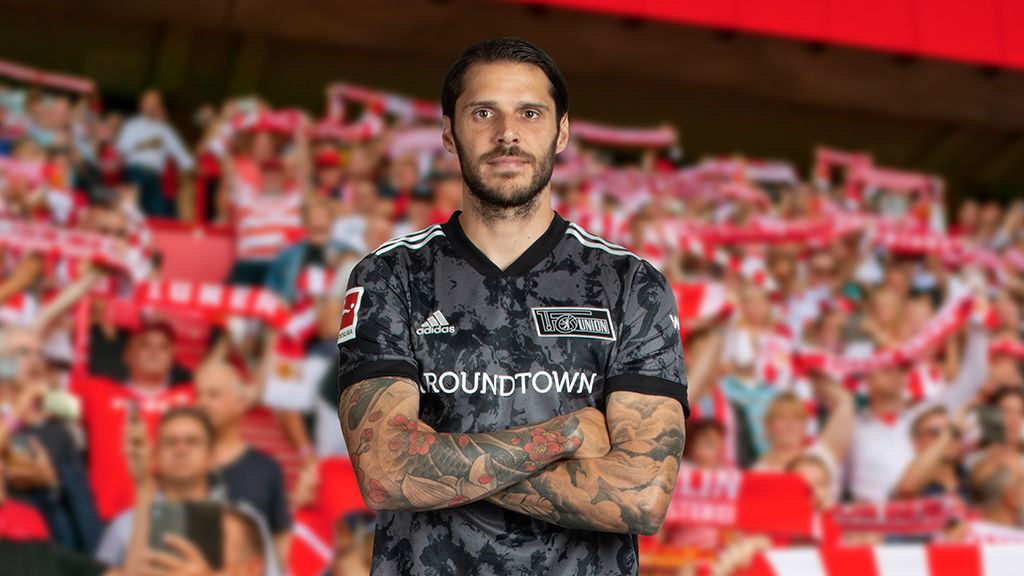 Ein Fußballspieler in einem schwarzen Trikot mit Tattoos steht mit verschränkten Armen vor einer jubelnden Menge im Hintergrund.