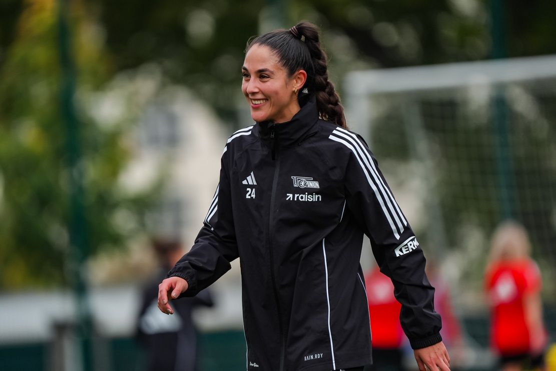 Lächelnde Fußballspielerin in schwarzer Trainingsjacke auf dem Spielfeld, umgeben von weiteren Sportlern.