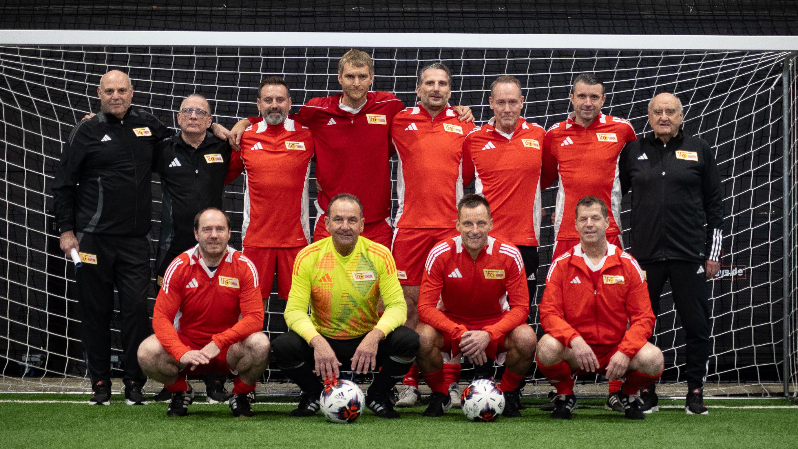 Gruppe von Fußballspielern in roten Trikots, stehend und kniend, vor einem Fußballtor in einer Sporthalle.