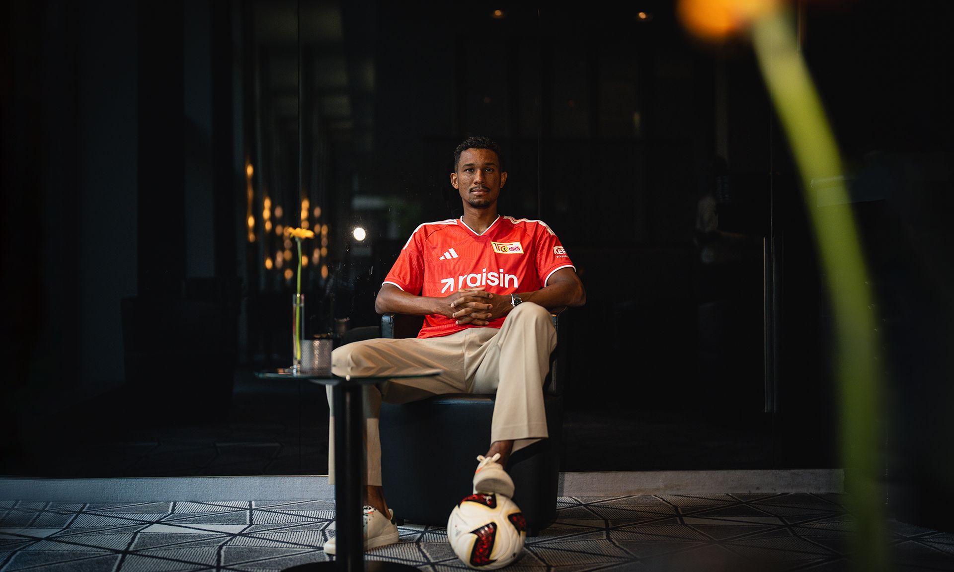 Ein Sportler sitzt entspannt auf einem Stuhl, trägt ein rotes Trikot und hat einen Fußball neben sich.