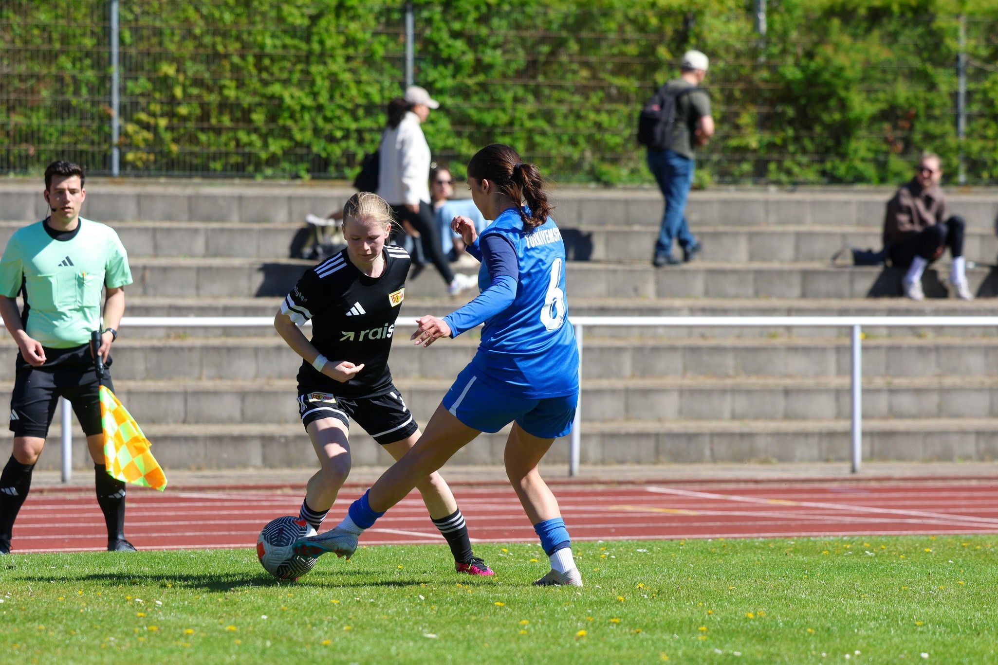 Zwei Fußballspielerinnen im Wettstreit um den Ball auf einem grünen Spielfeld, Zuschauer im Hintergrund.