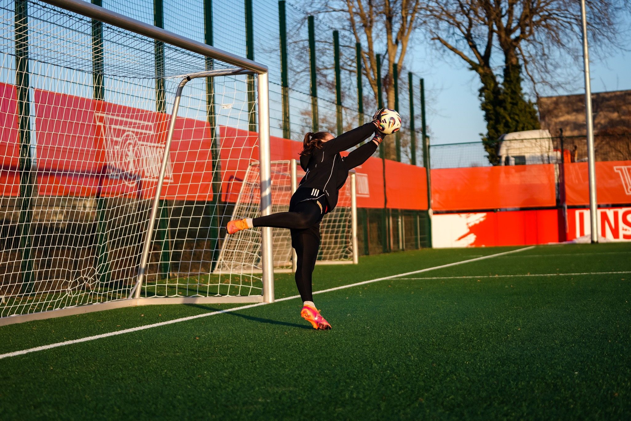 Eine Torhüterin fängt einen Ball in einer Fußballspielumgebung auf einem Kunstrasenplatz.