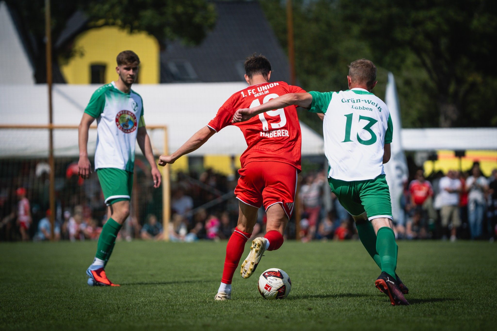 Ein Fußballspieler in rotem Trikot dribbelt den Ball, während zwei Gegner in grünen Trikots versuchen, ihn zu stoppen.