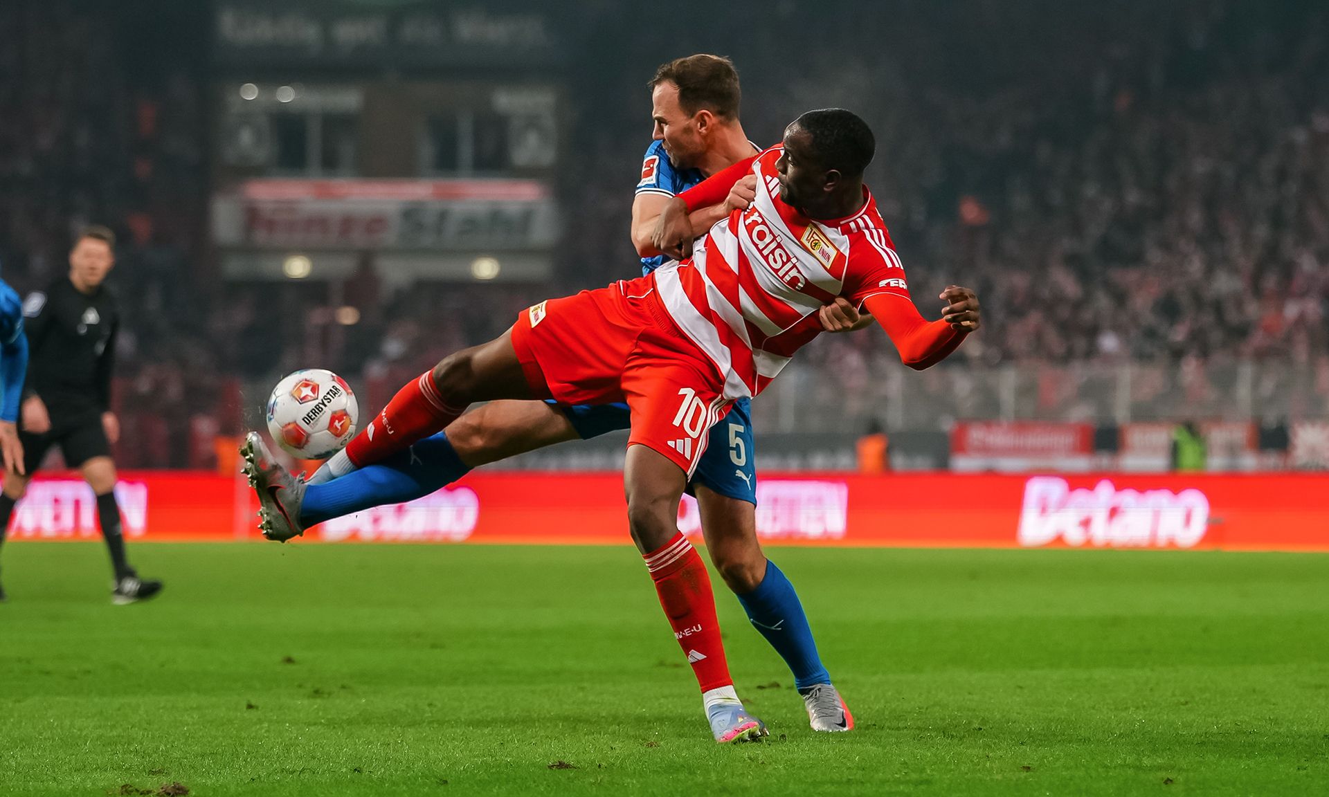 Ein Fußballspieler in roter und weißer Uniform wird von einem Gegner in blauer Uniform in einem Zweikampf umgeworfen.