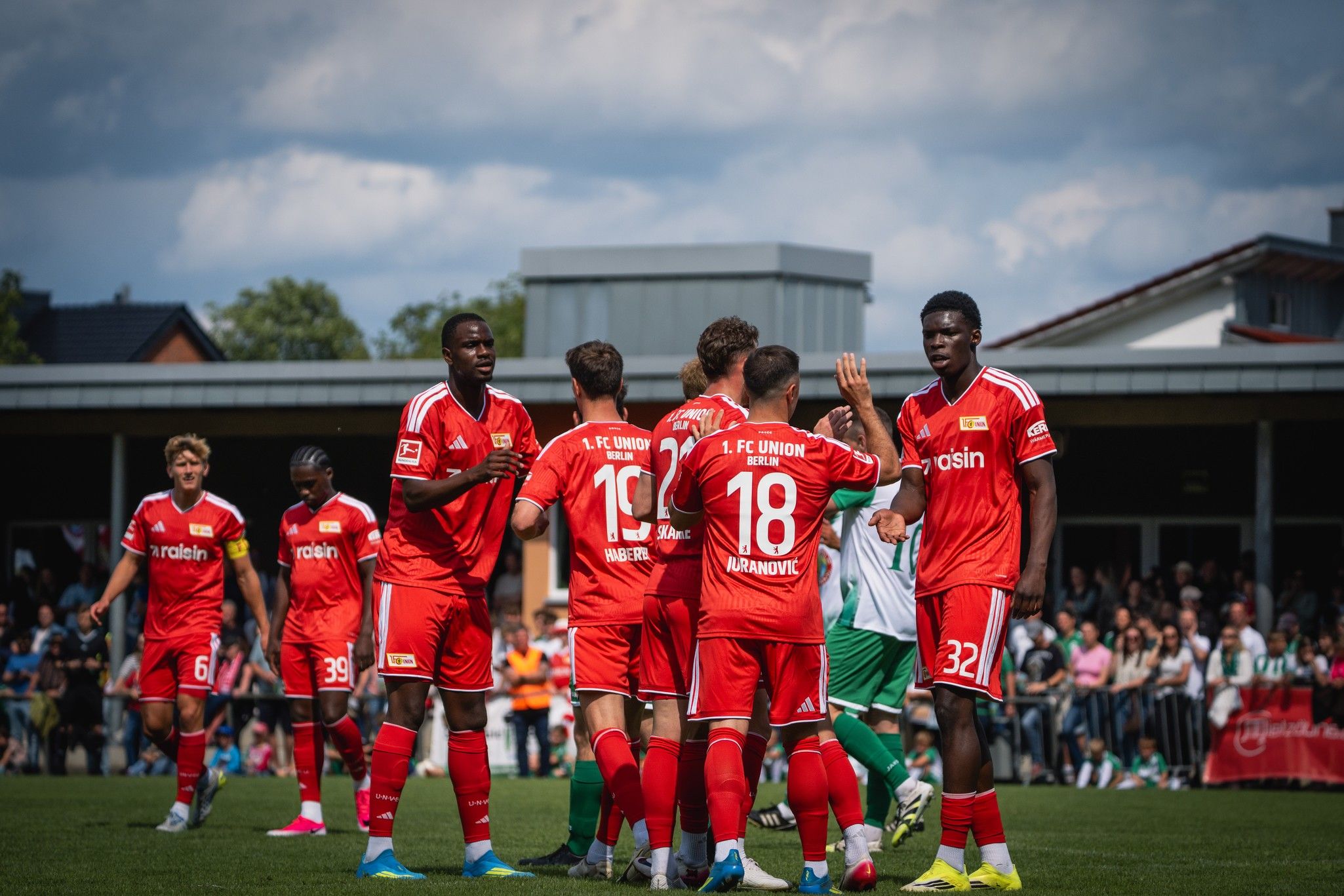 Spielszene einer Fußballmannschaft in roten Trikots, die sich um einen Spieler versammelt. Zuschauer im Hintergrund.