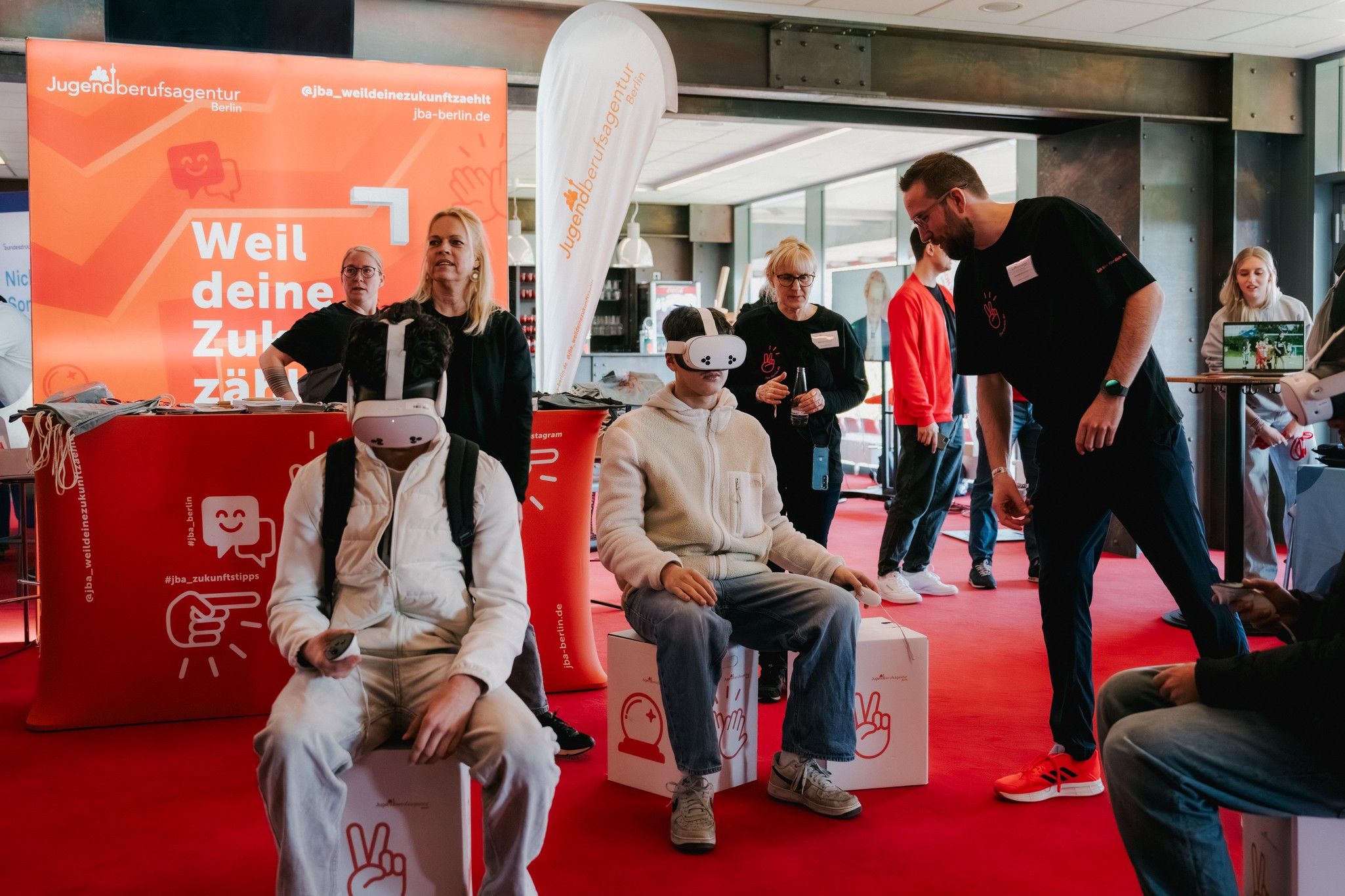 Zwei Personen mit VR-Headsets sitzen auf weißen Kisten in einem modernen Raum, umgeben von Besuchern und Informationsständen.