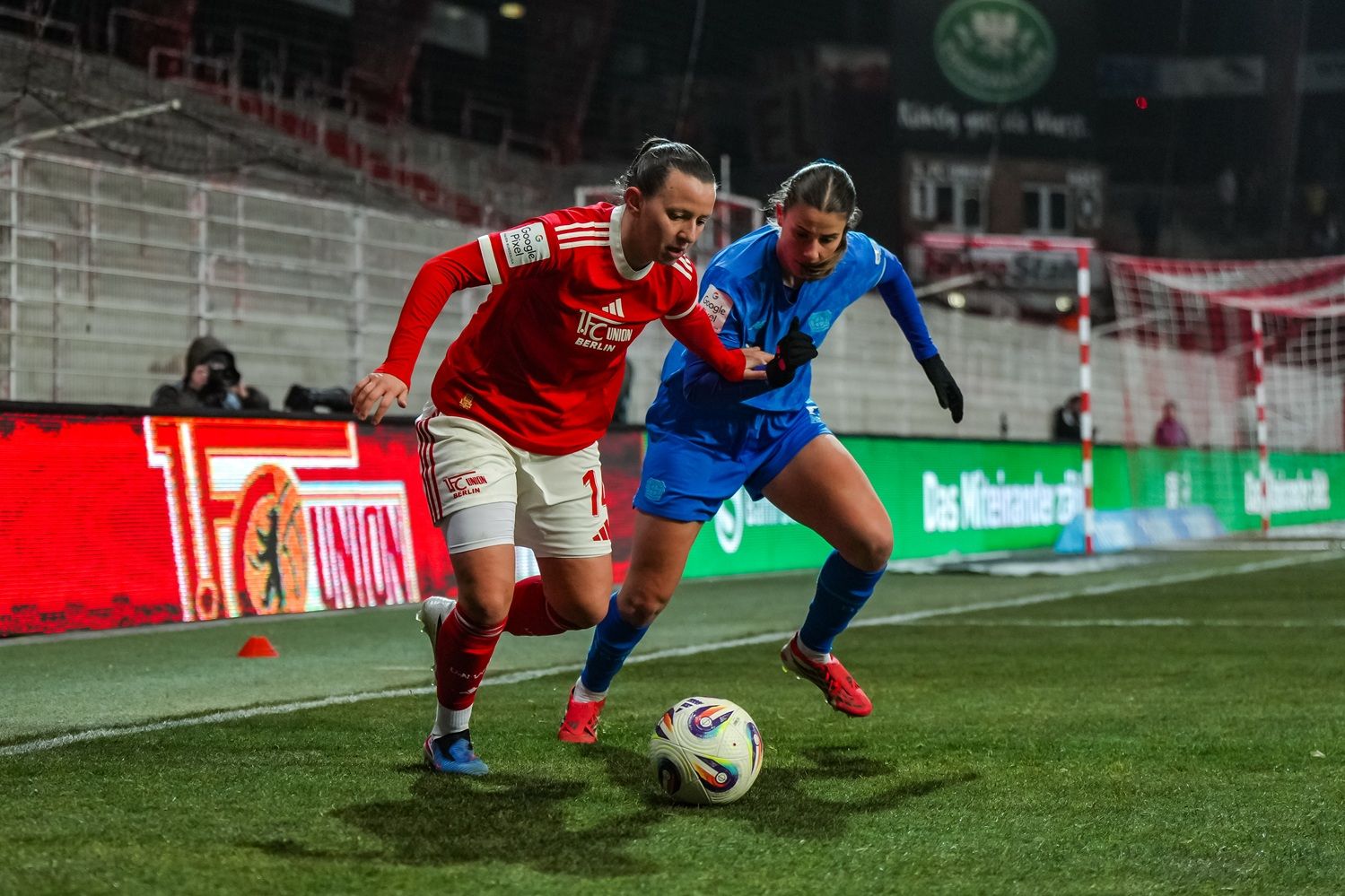 Zwei Fußballspielerinnen kämpfen um den Ball auf dem Spielfeld während eines Spiels unter Flutlicht.