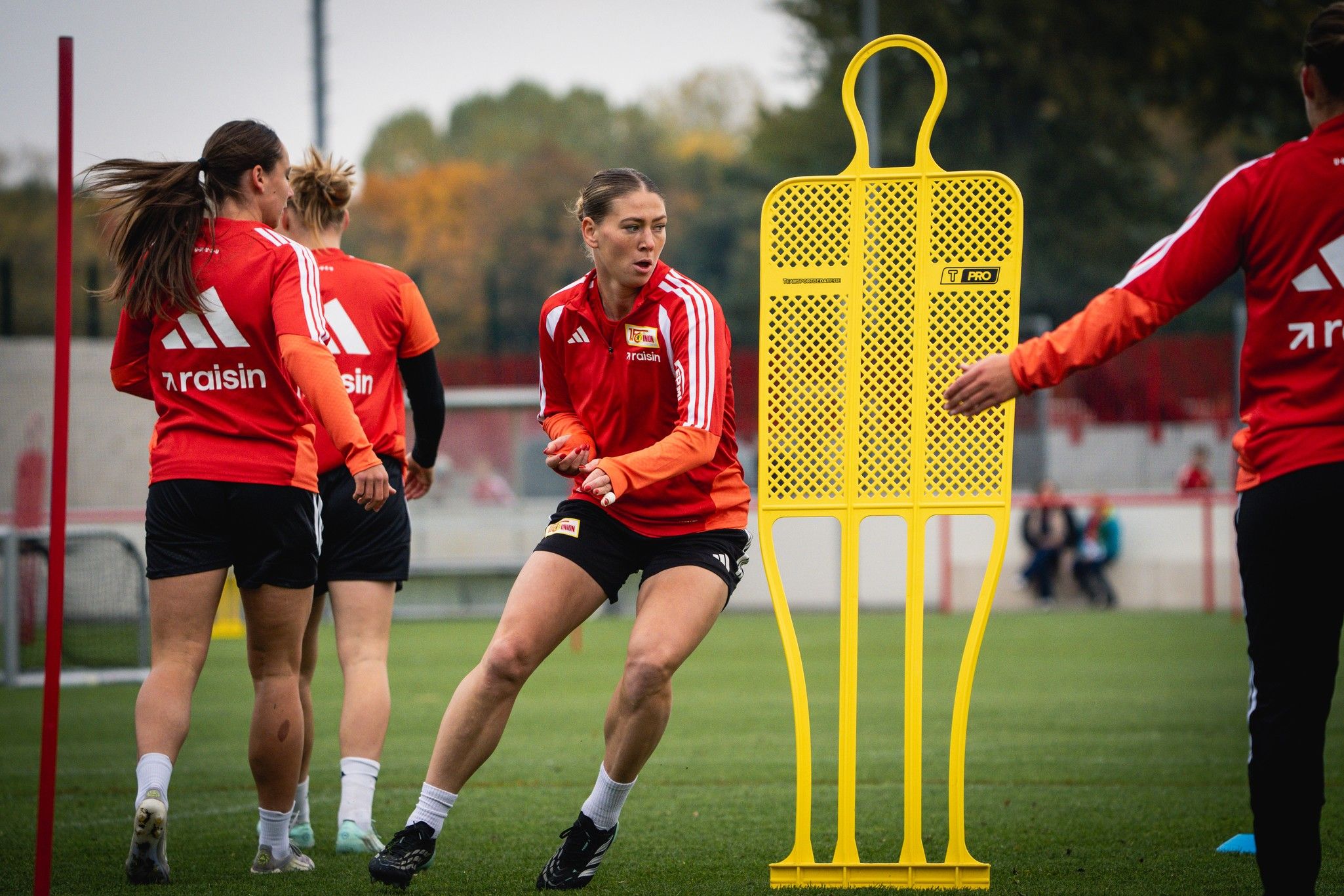 Frau in Sportkleidung trainiert auf einem Fußballplatz, während zwei weitere Spielerinnen im Hintergrund an einem Dummy vorbeilaufen.
