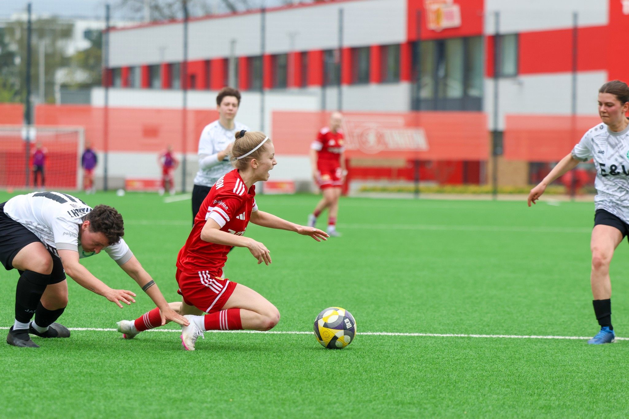 Eine Fußballspielerin in rotem Trikot fällt auf den Kunstrasen, während zwei gegnerische Spielerinnen auf den Ball zusteuern.