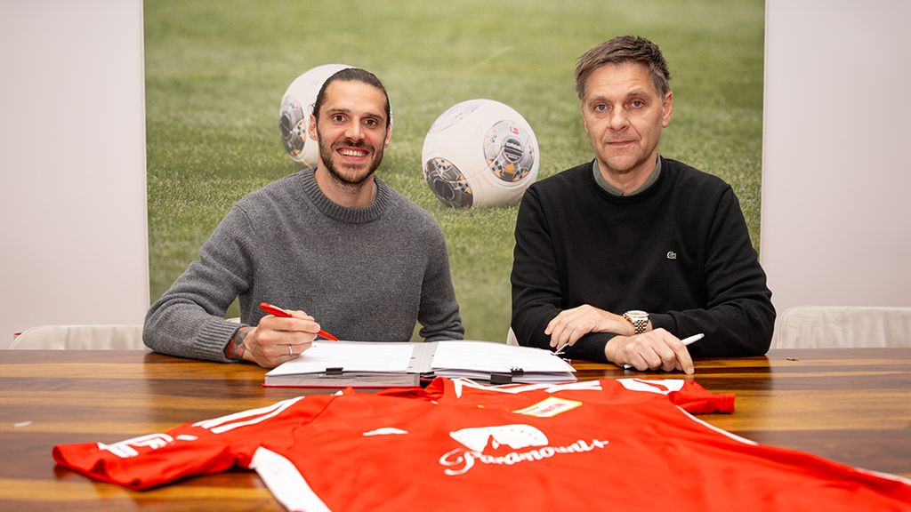 Zwei Männer sitzen an einem Tisch, einer unterschreibt einen Vertrag. Auf dem Tisch liegt ein rotes Fußballtrikot.