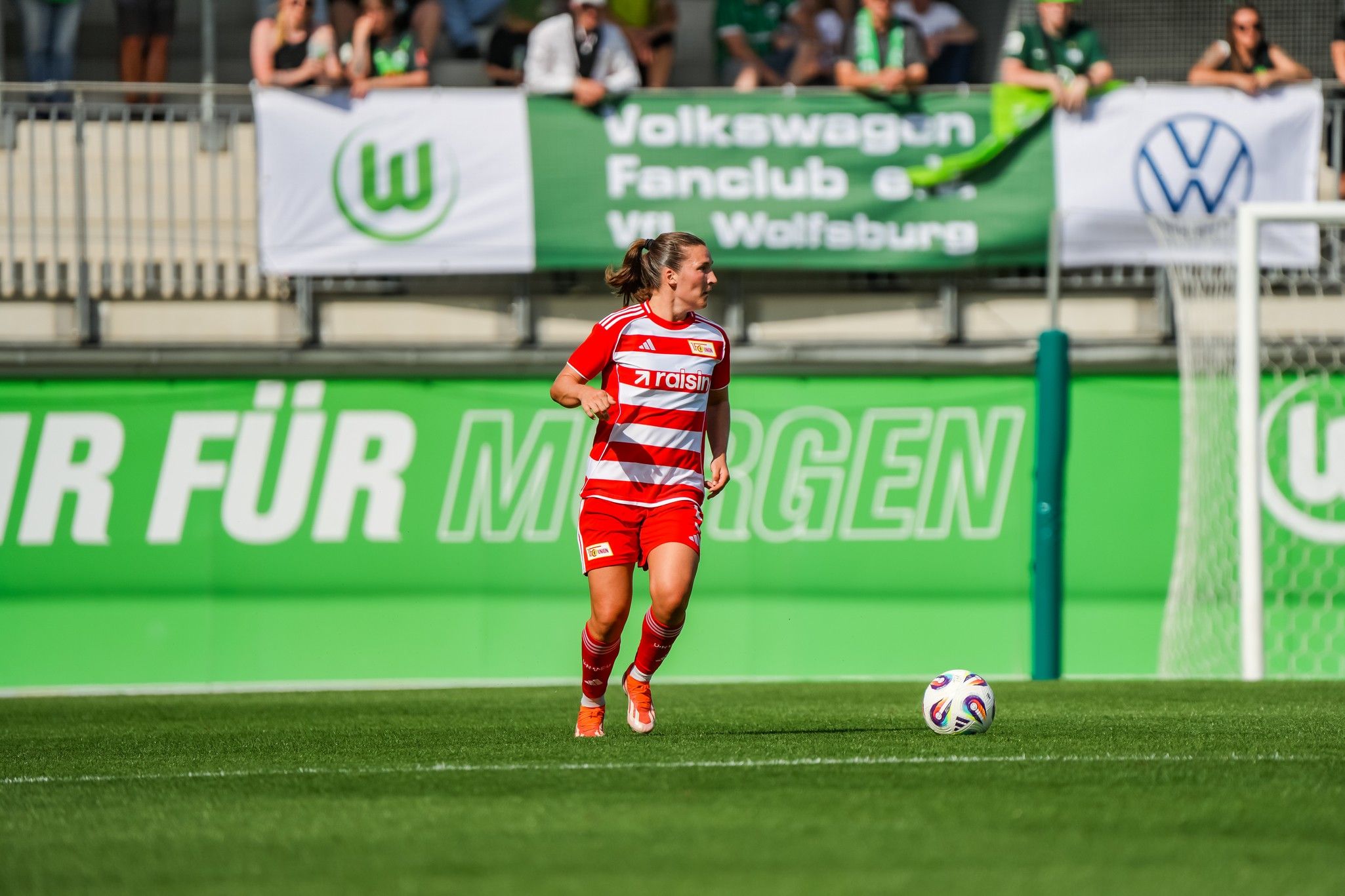 Eine Fußballspielerin in rotem Trikot rennt über das Spielfeld, im Hintergrund sind Fans und Banner zu sehen.