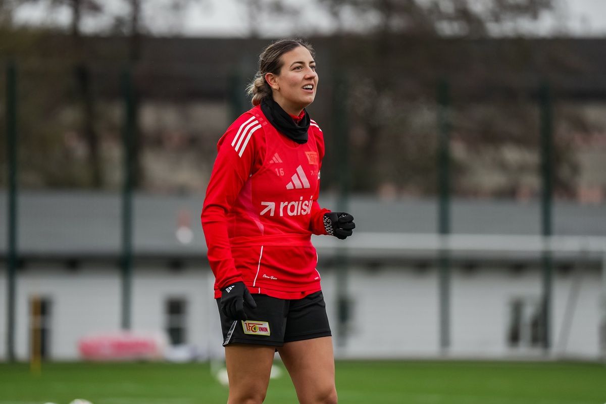 Sportlerin in roter Trainingsjacke und schwarzem Schal, steht auf einem Fußballplatz und ist mit einem Lächeln am Übungen beteiligt.