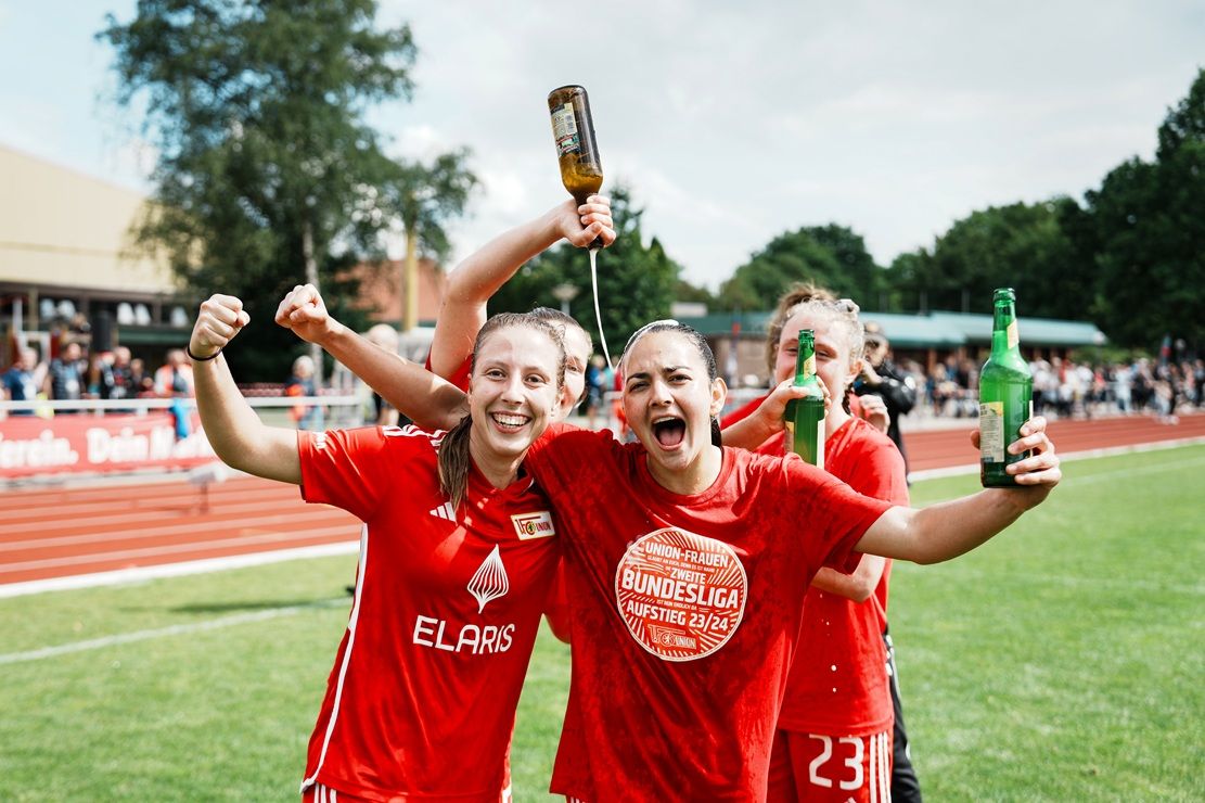 Zwei freudige Fußballspielerinnen in roten Trikots feiern mit Getränken auf dem Platz, umgeben von weiteren Teamkolleginnen.
