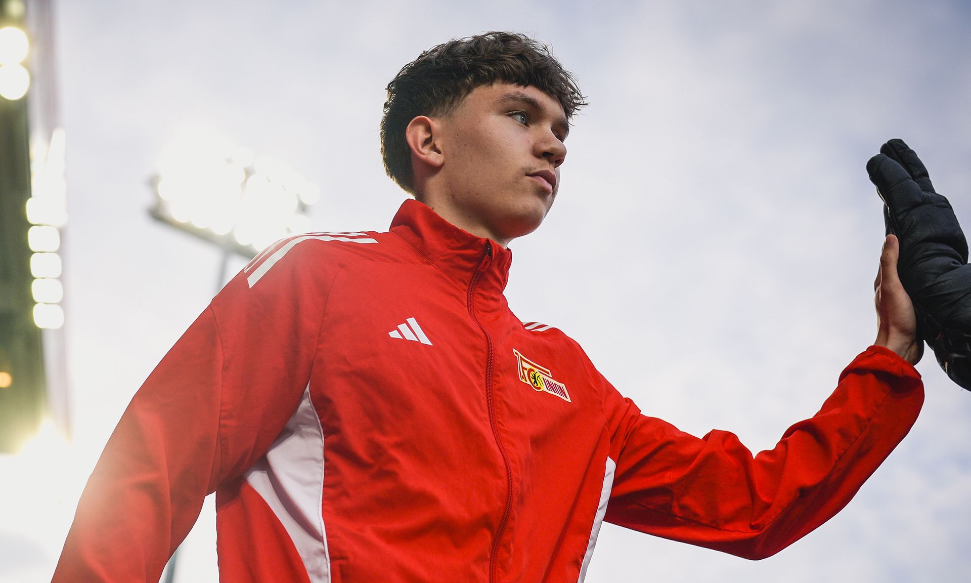 Ein Sportler in einem roten Trainingsanzug mit weißen Details, der auf einem Stadionfeld steht und seine Hand hebt.