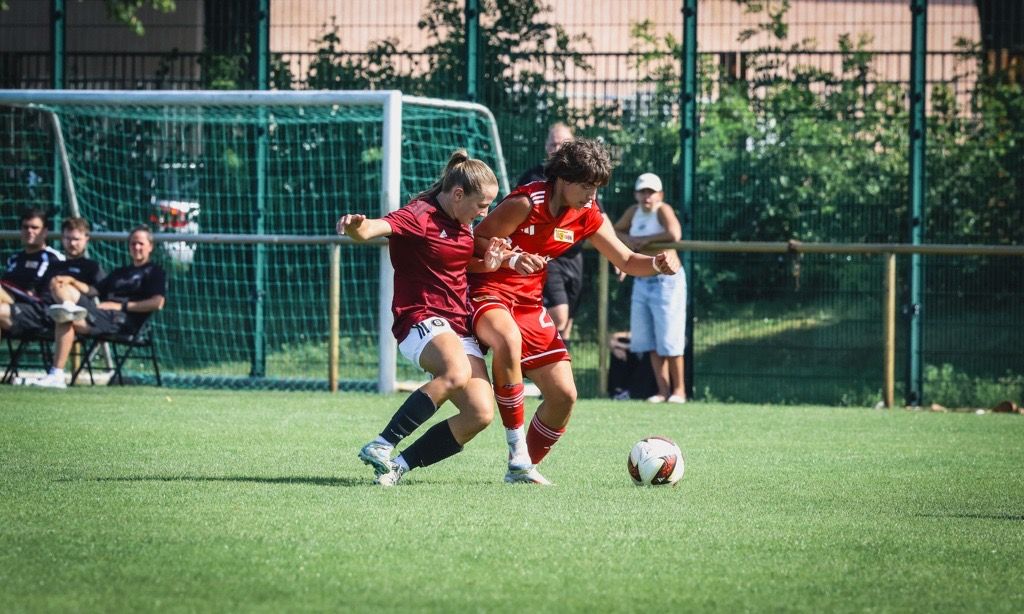 Zwei junge Fußballspieler im Wettkampf um den Ball auf einem Grünfeld, im Hintergrund Zuschauer und ein Tor.
