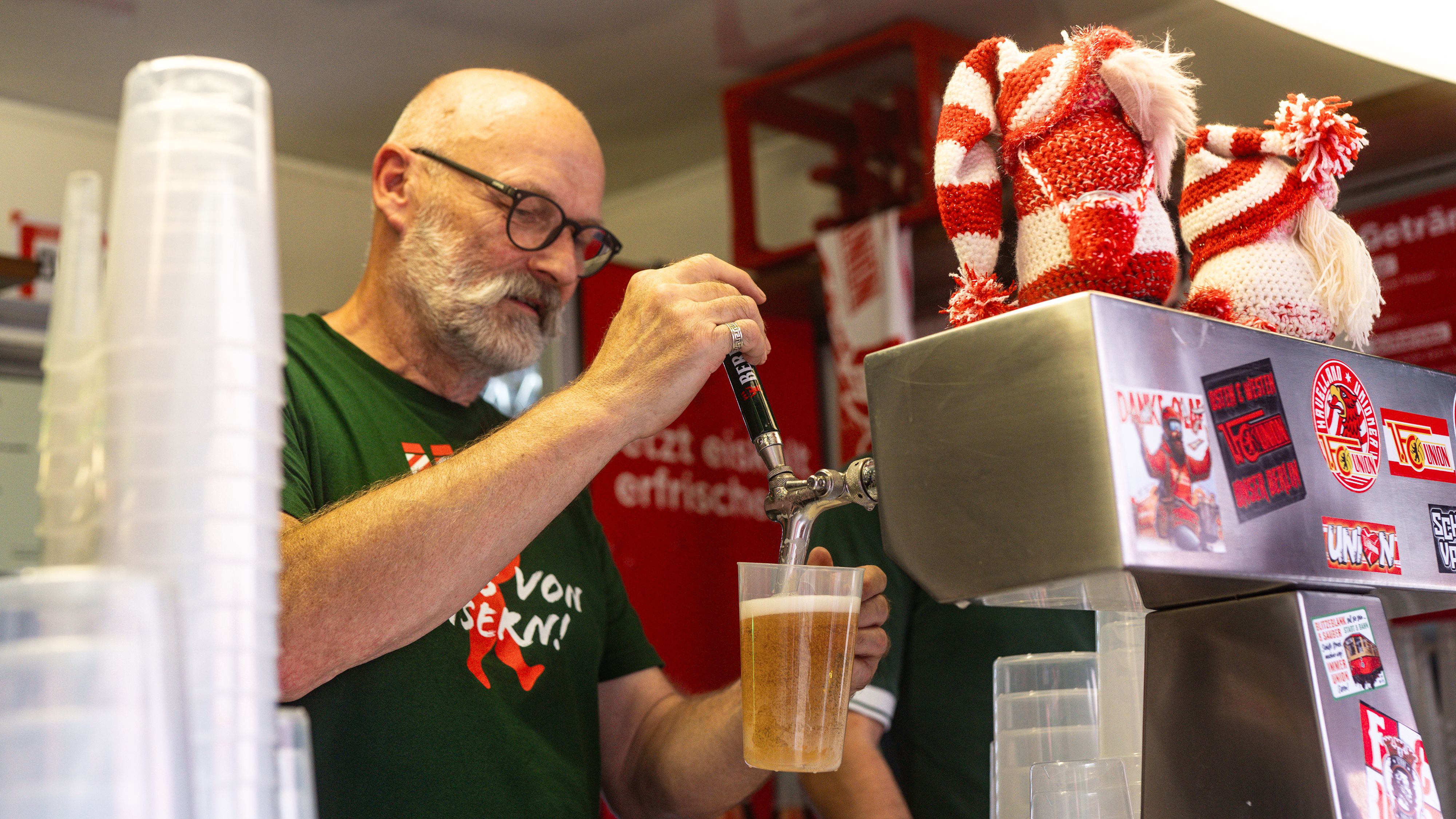 Ein Mann mit Brille zapft Bier in ein Plastikbecher an einem Getränkestand. Er trägt ein grünes Shirt.
