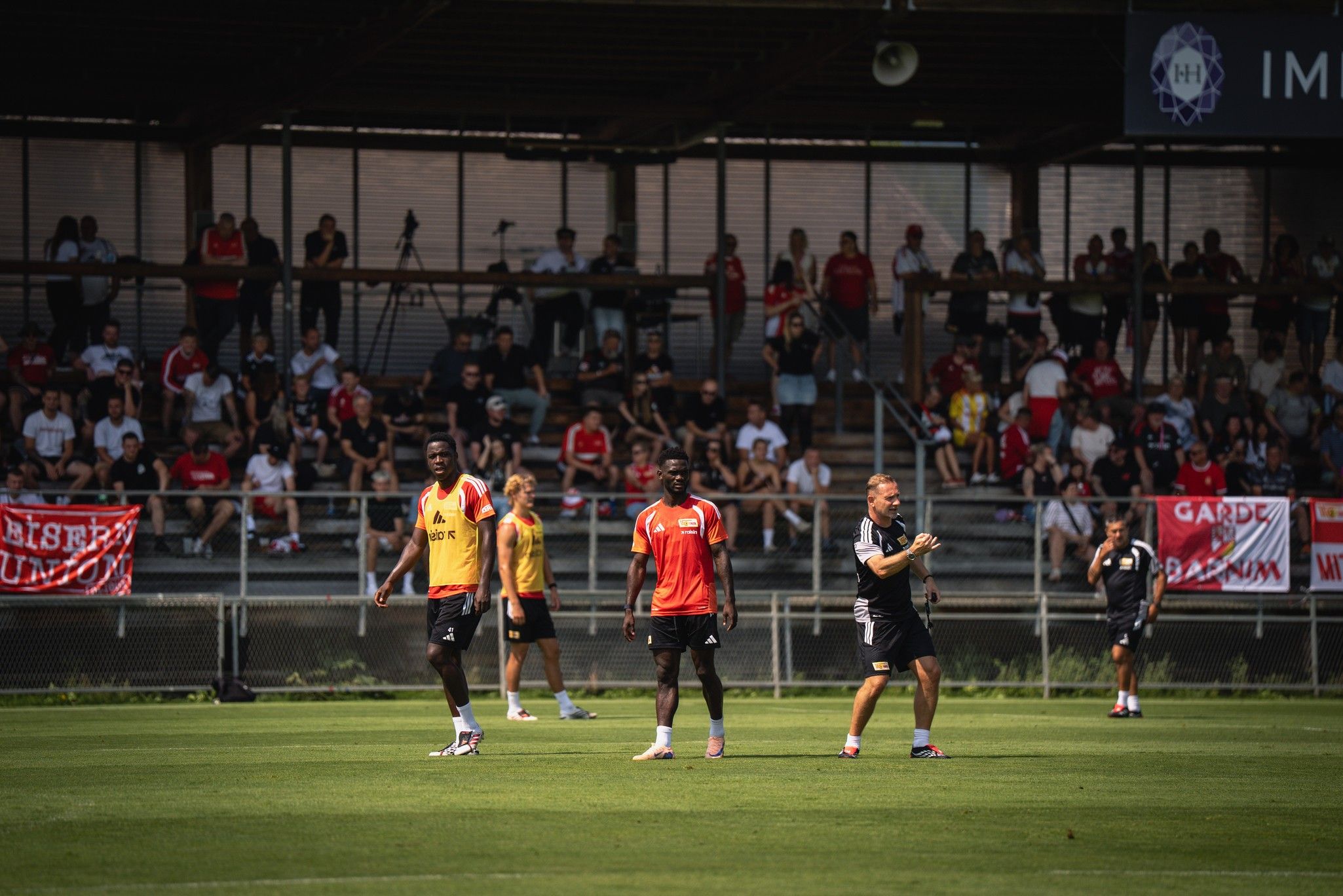 Fußballtraining auf einem Platz, Spieler in roten und gelben Trikots sowie ein Trainer, Fans im Hintergrund.